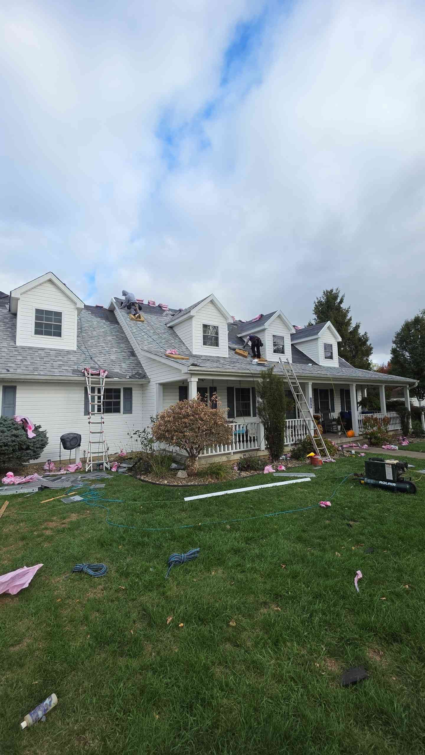House with roof damage, debris scattered on lawn under cloudy sky.