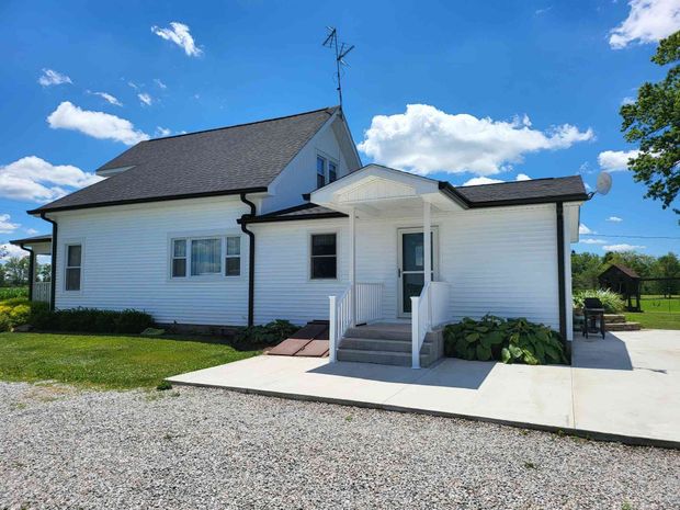 White farmhouse with dark roof, porch, and concrete patio against a blue sky with clouds.