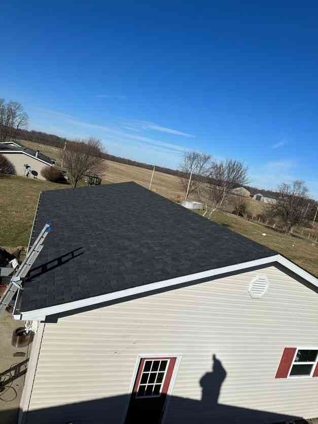 Newly shingled roof on a tan house with white trim; ladder leans against the roof; blue sky.