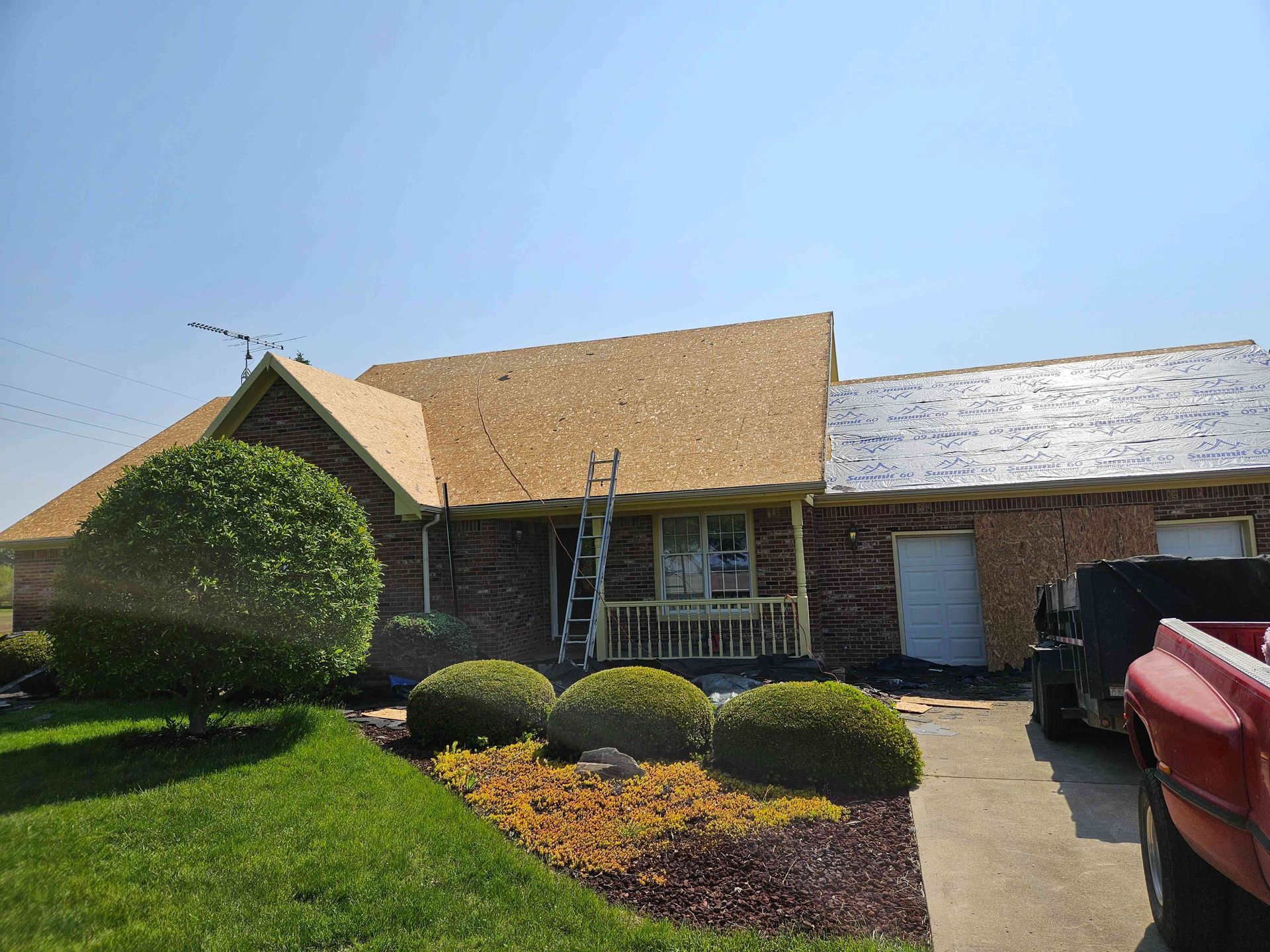 House with roof partially covered in new shingles. Workers and ladder present. Landscaping in foreground.