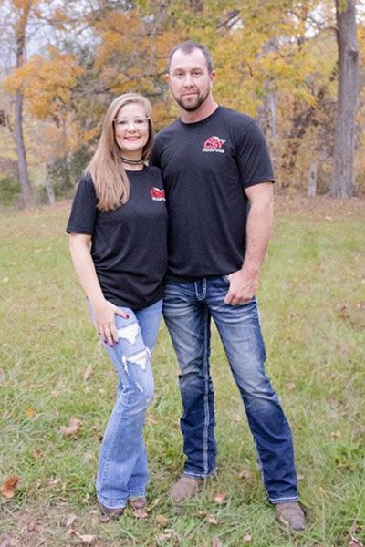 Two people in black shirts and jeans standing together in a grassy park with autumn trees.