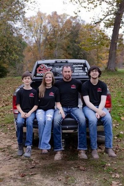 Four people sitting on the tailgate of a pickup truck in a wooded outdoor setting.