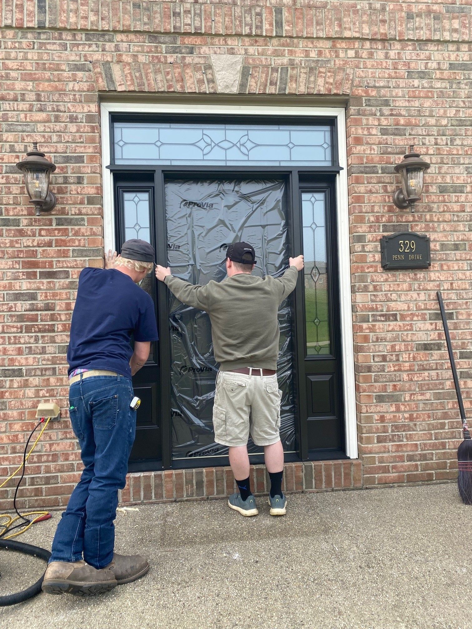Two men are working on a front door of a brick house.