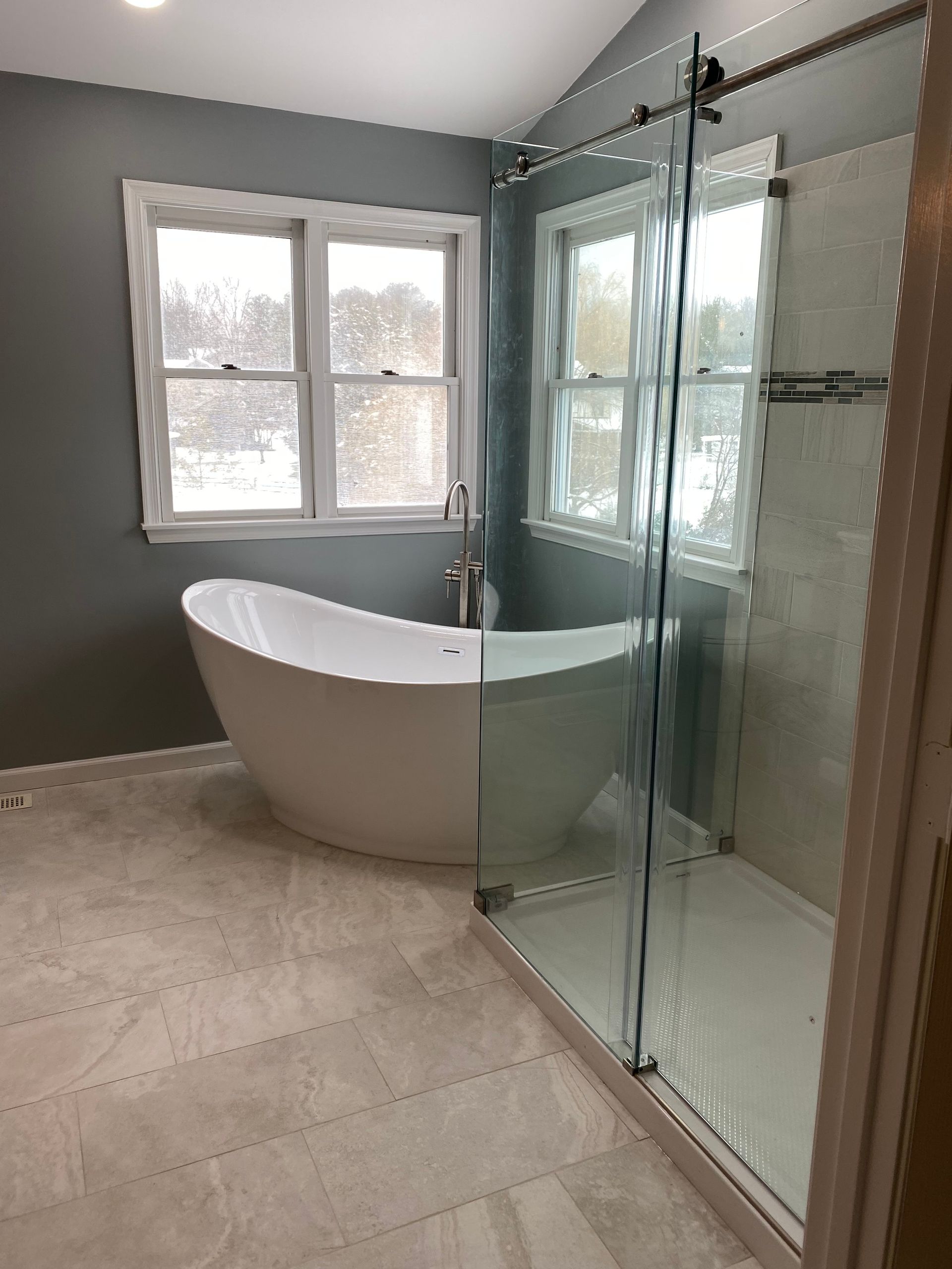 Modern bathroom with freestanding tub, glass shower enclosure, and two windows on gray walls
