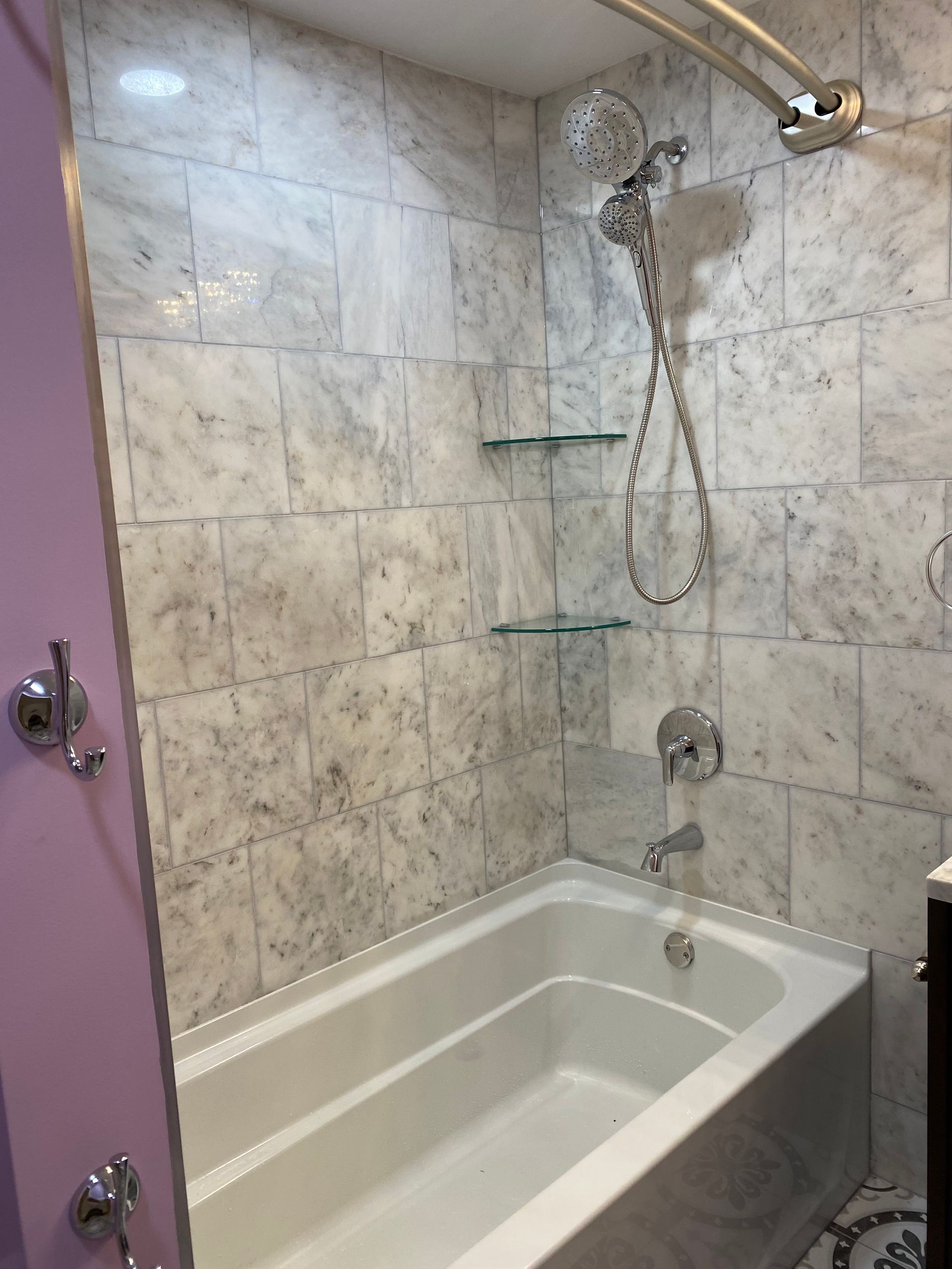 Small tiled bathroom shower with glass corner shelves, handheld showerhead, and white tub with beige tile walls