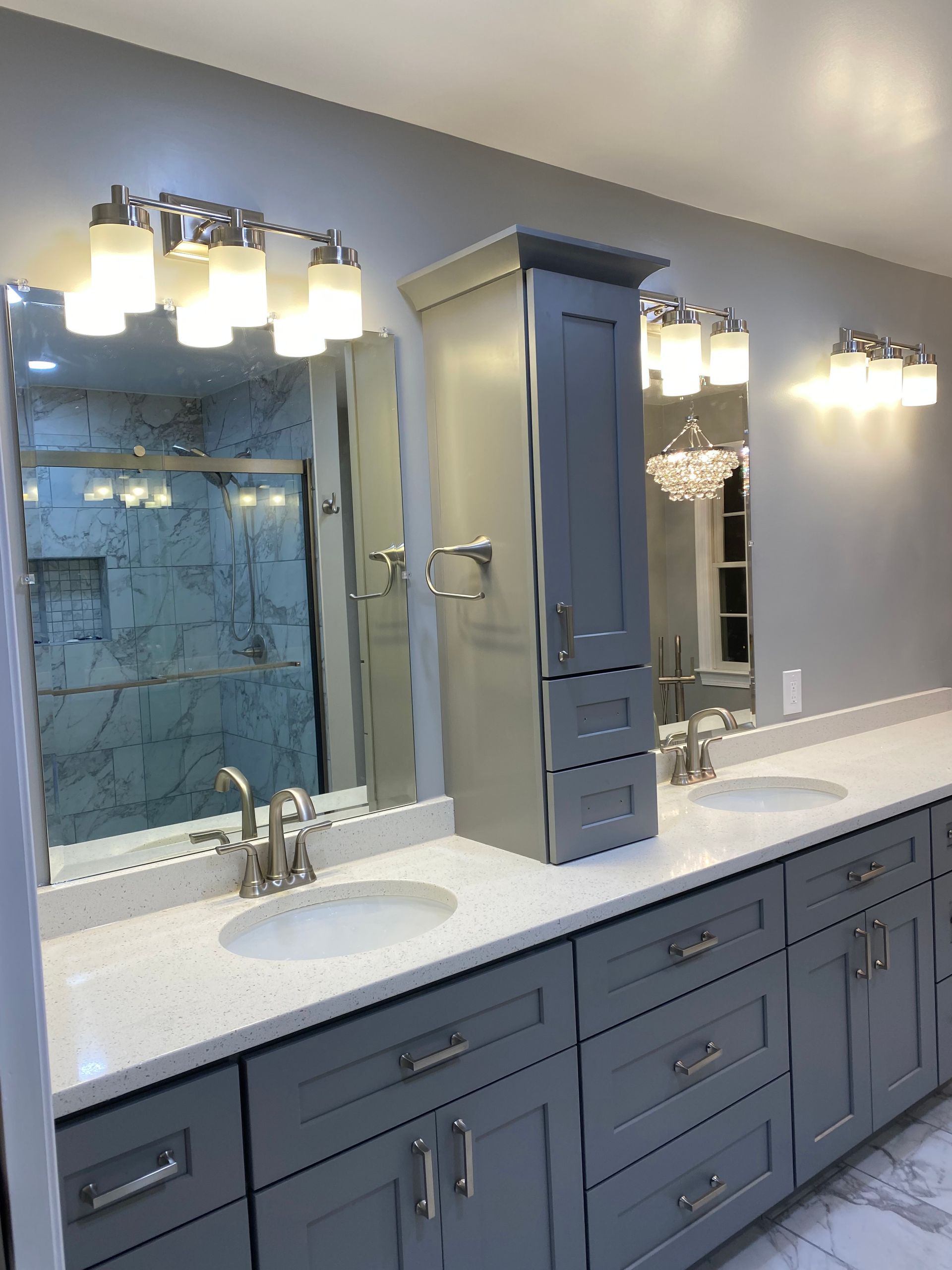 Modern bathroom vanity with two sinks, mirrors, sconces, and a tall gray cabinet on a white countertop.