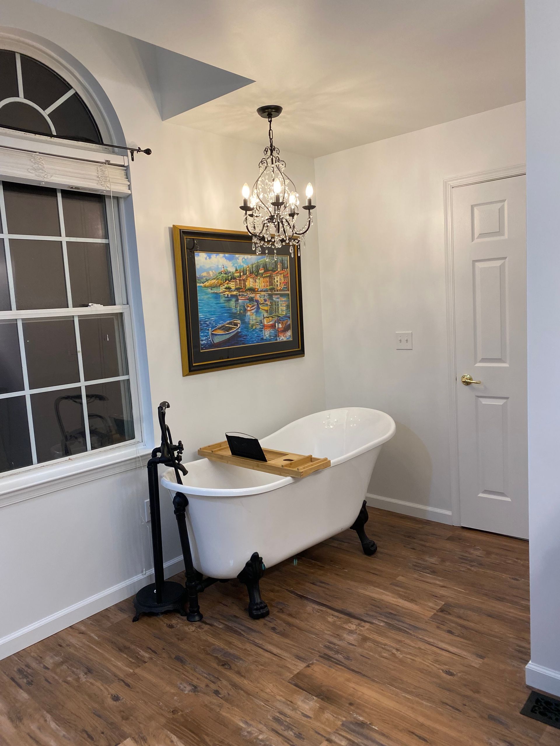 Small room with a white bathtub, chandelier, framed painting, and window with blinds and curtain