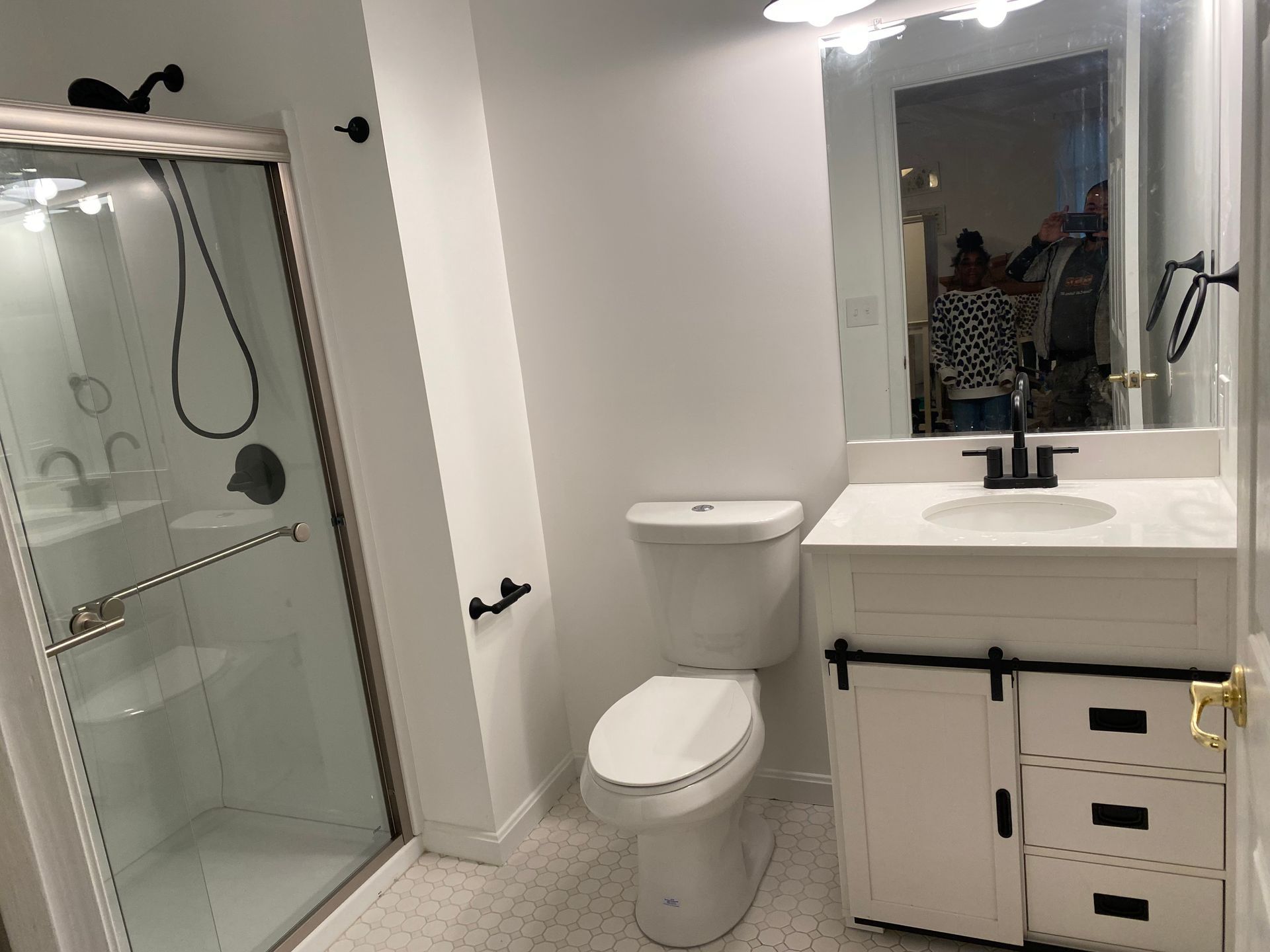 Small bathroom with shower, toilet, and white vanity with black hardware and mirror.