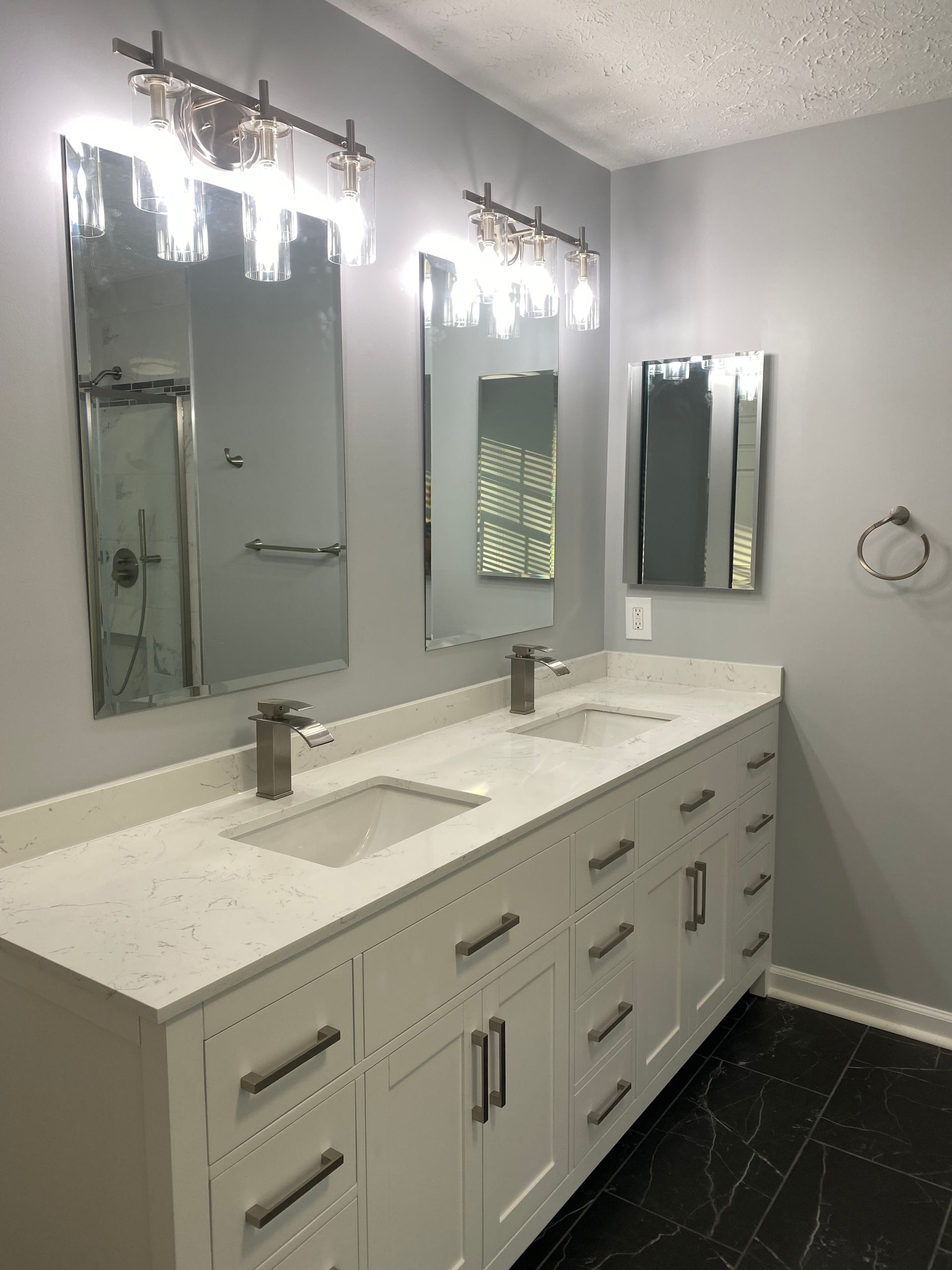Modern double-sink bathroom vanity with large mirrors, bright lights, white countertop, and dark tile floor