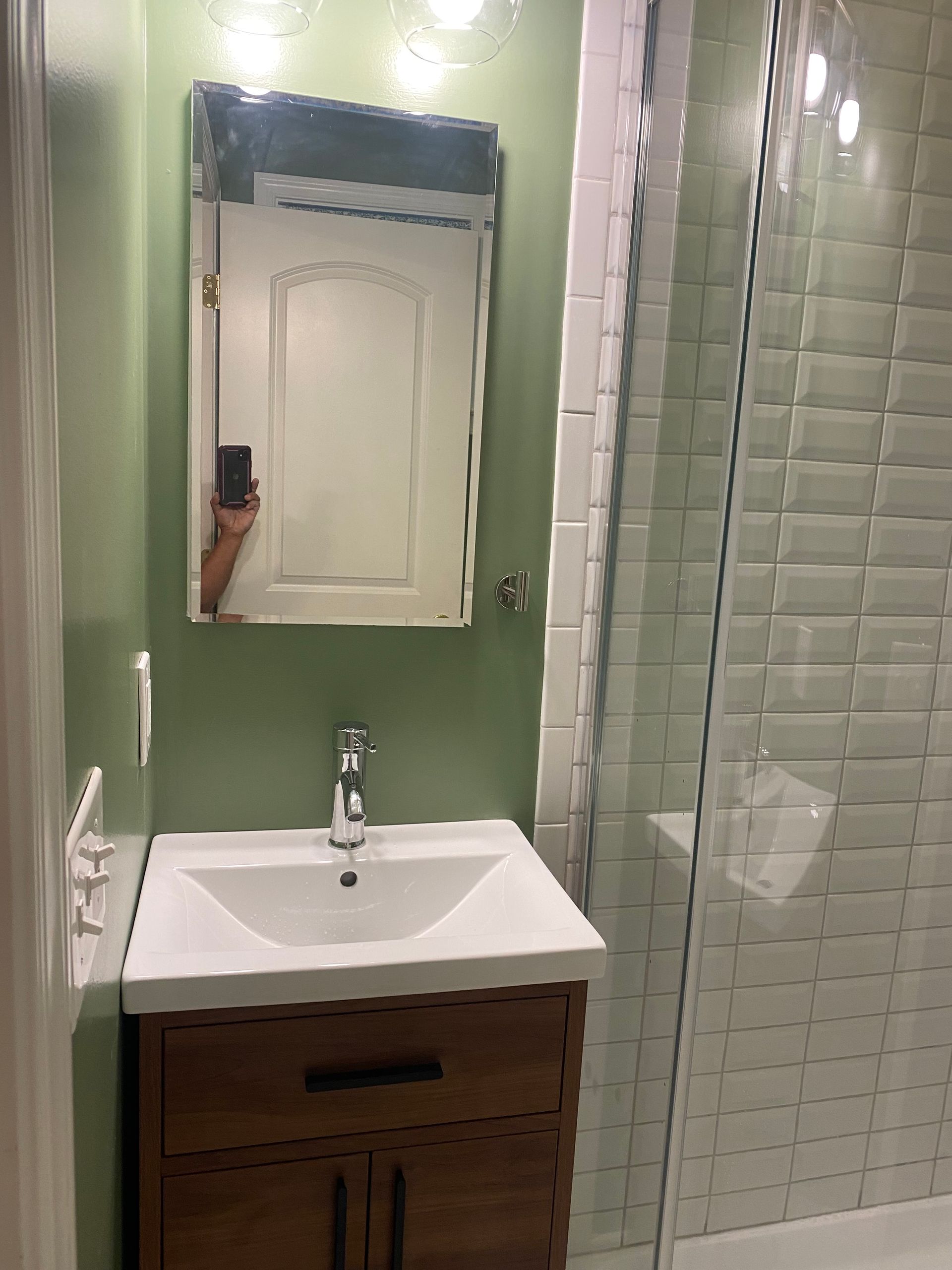 Small bathroom vanity with sink, mirror cabinet, and green tiled walls; phone visible in mirror.