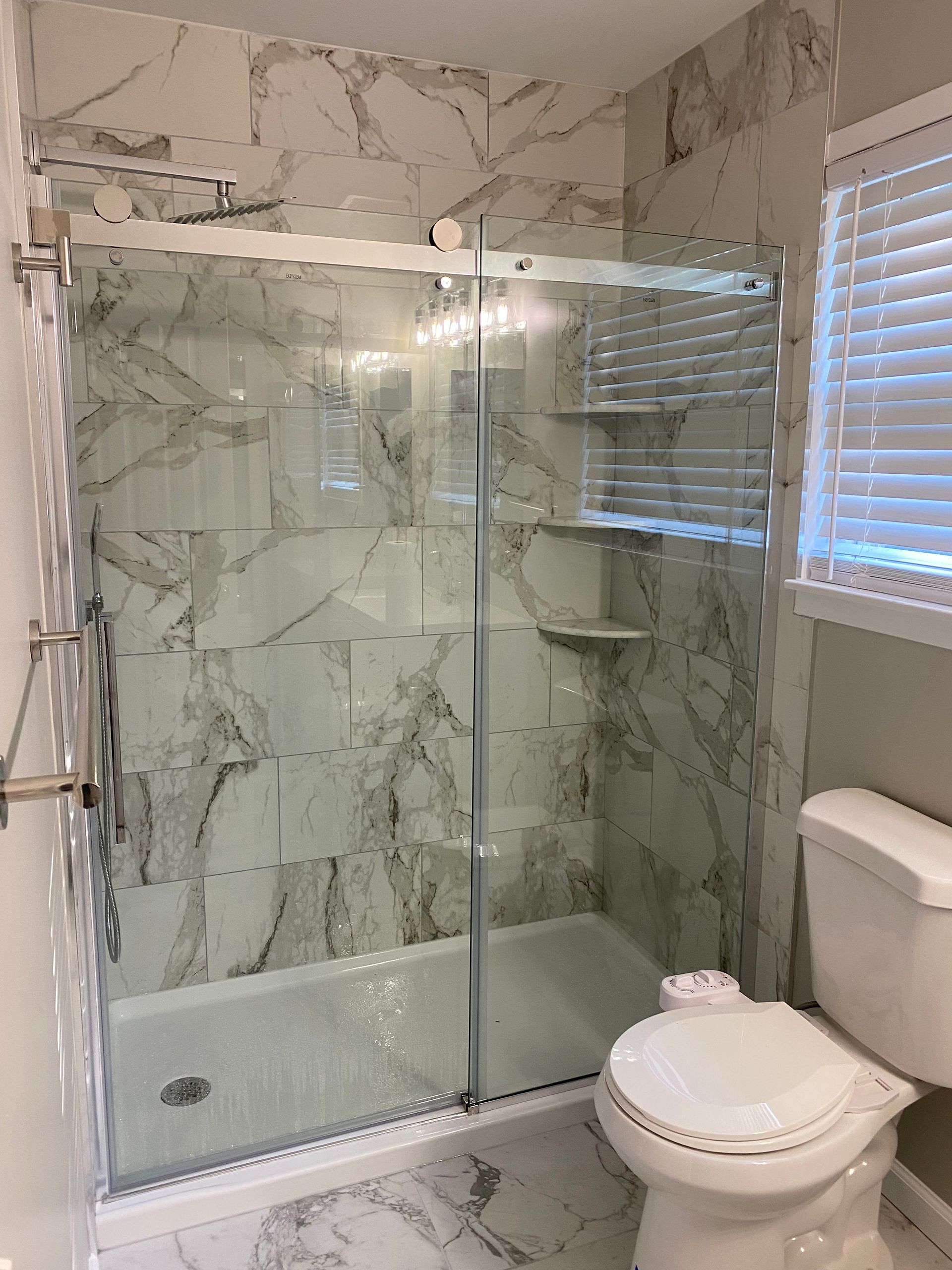 Modern bathroom with marble-tile shower enclosure, glass doors, and toilet next to a window with blinds