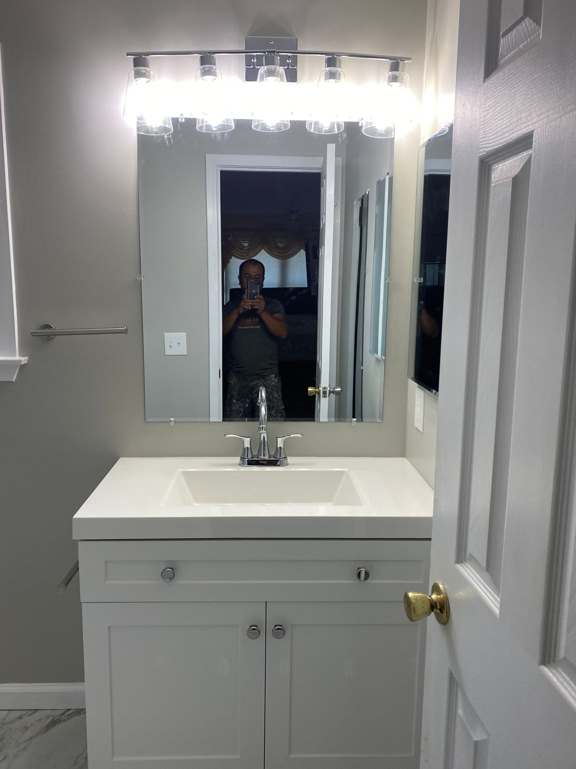 Small white bathroom with vanity, sink, mirror lights, and a person reflected in the mirror