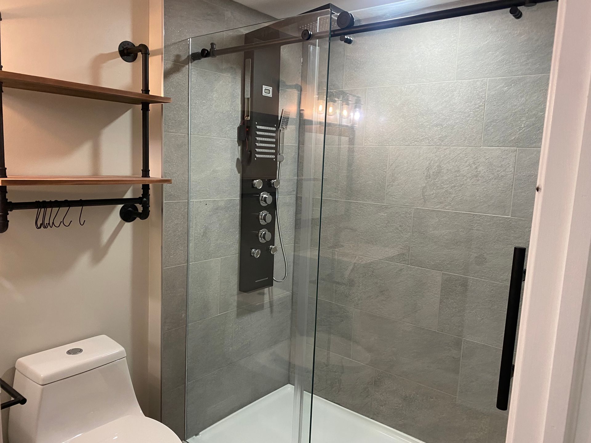 Modern bathroom with a glass shower enclosure, black shower panel, white toilet, and wood shelves.