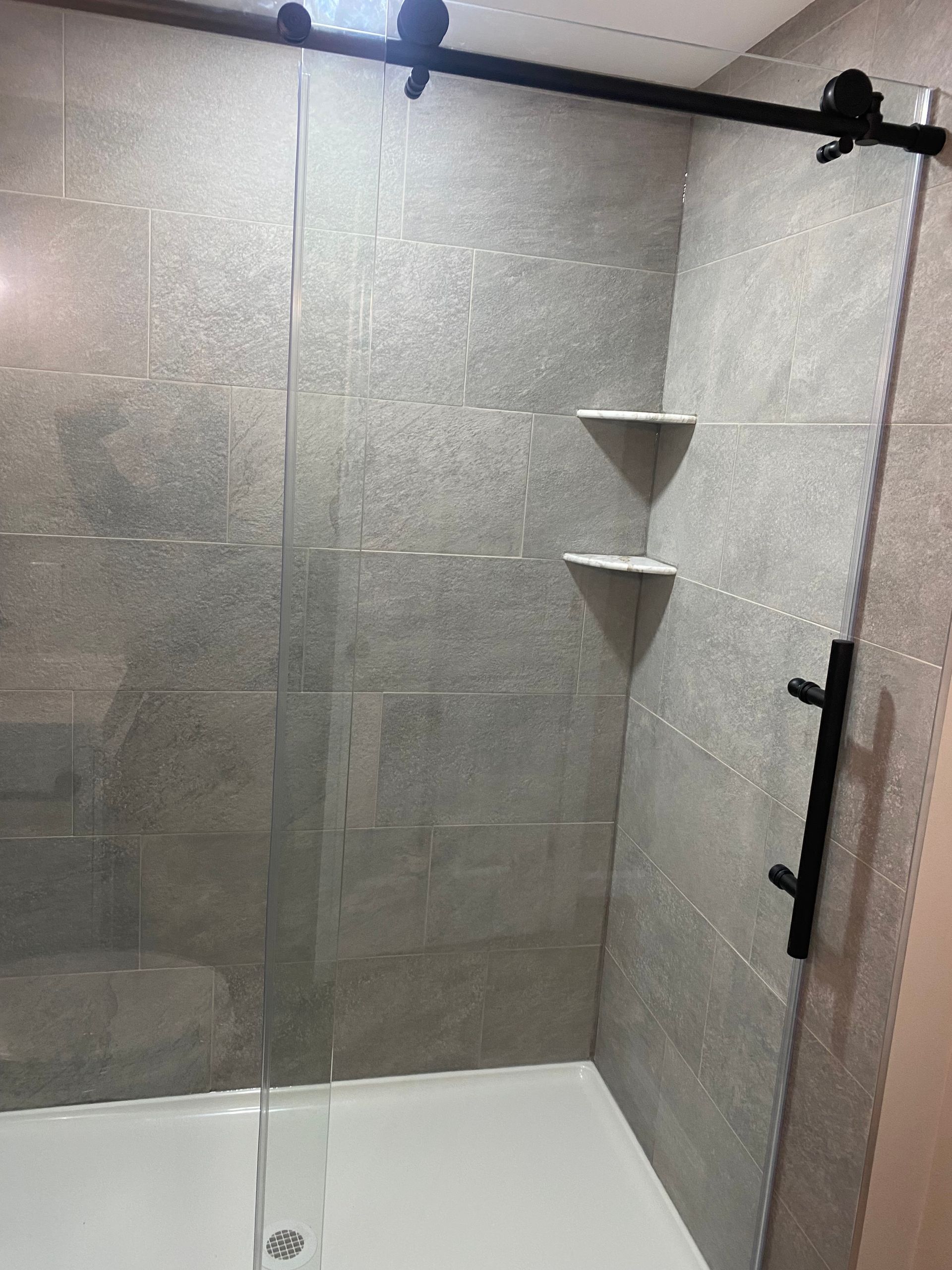 Glass-enclosed shower with gray tile walls, white base, black hardware, and two corner shelves