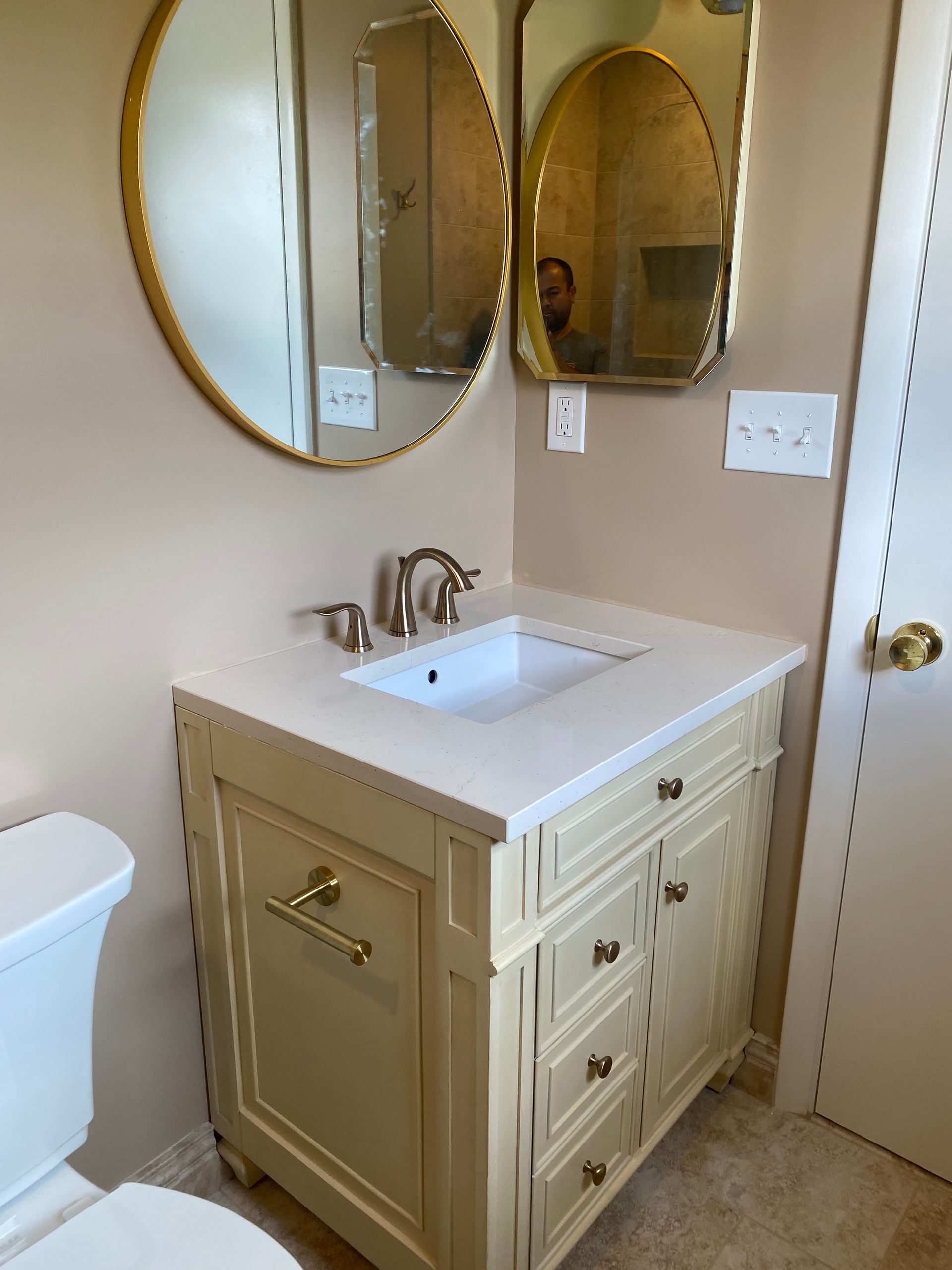 Beige bathroom vanity with sink, gold mirror, and white door against tan walls.