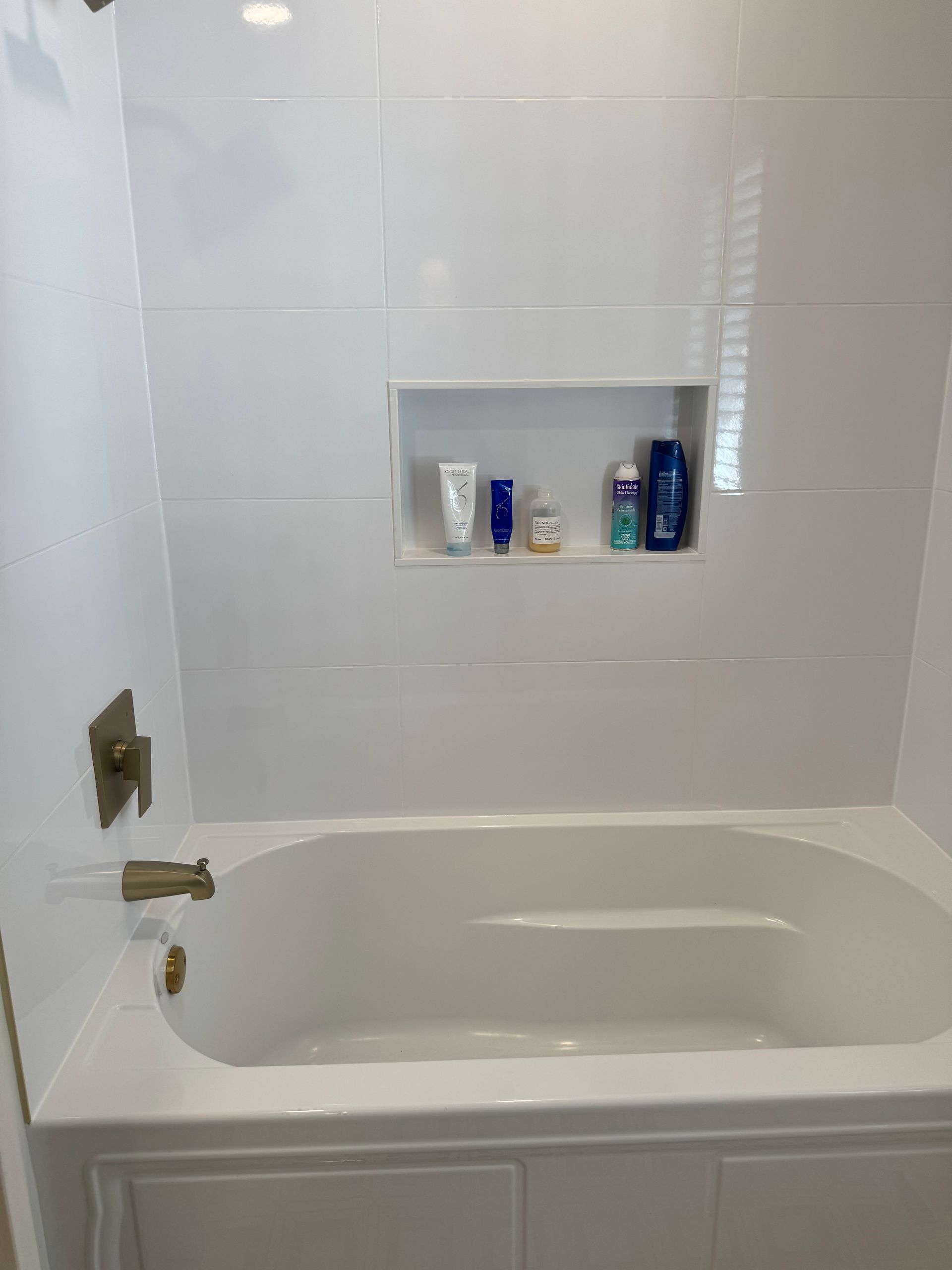 White tiled bathroom with a built-in bathtub, chrome fixtures, and a recessed shelf with toiletries.