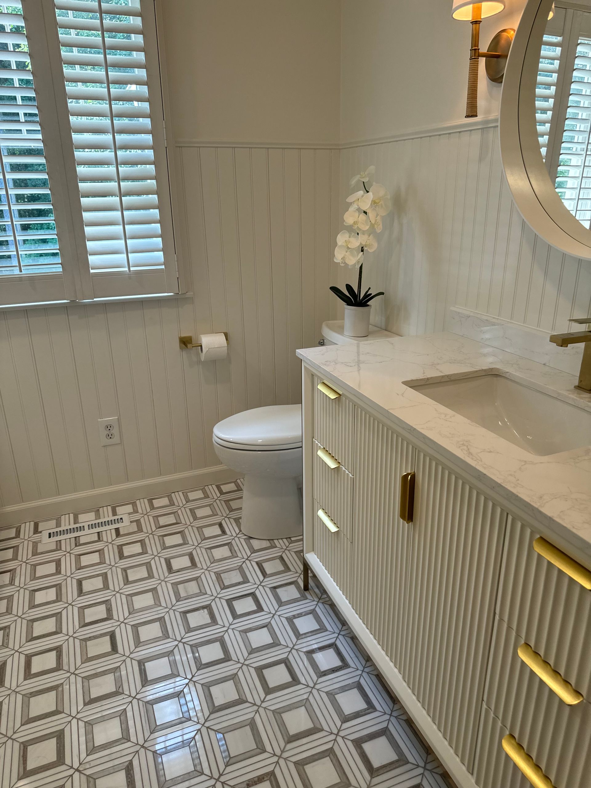 Modern bathroom with patterned tile floor, vanity sink, white toilet, and window shutters.