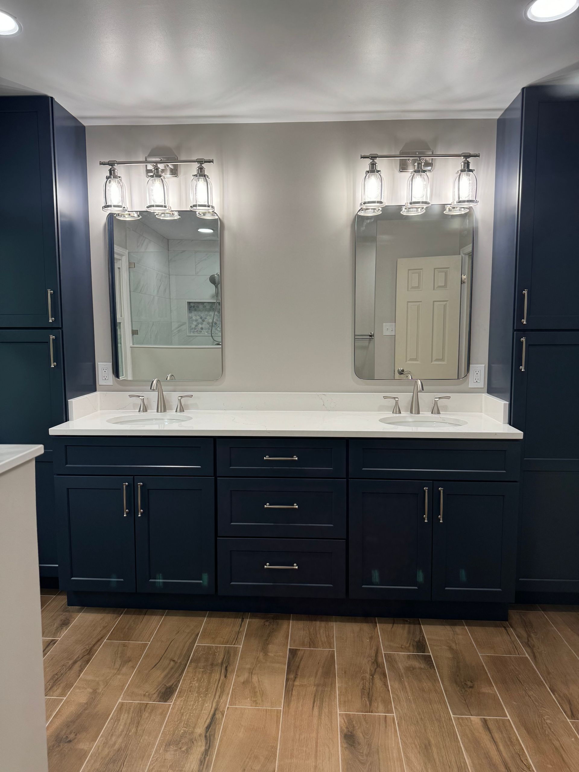 Bathroom vanity with dark blue cabinets, double sinks, mirrors, and wood-look flooring