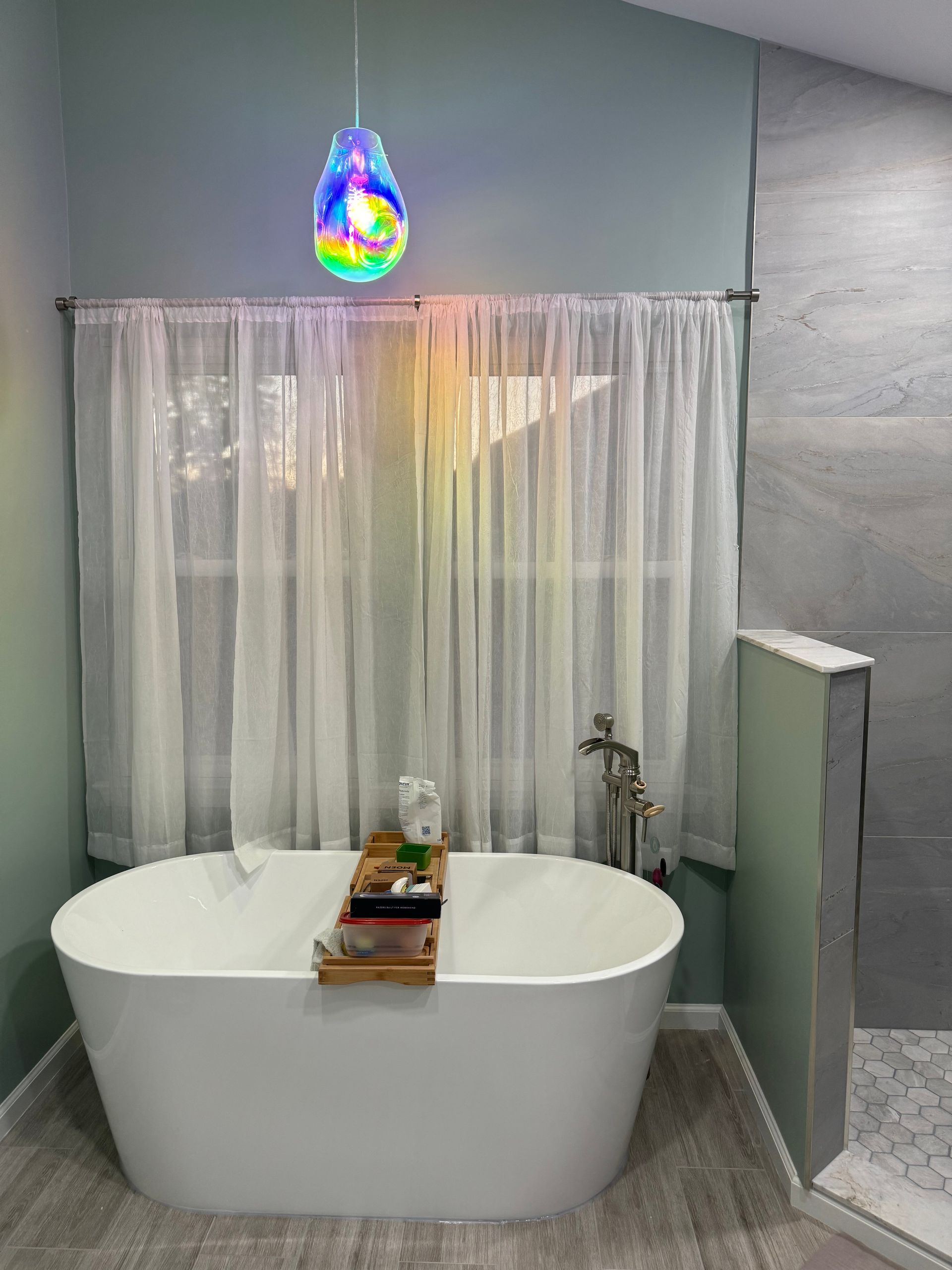 Modern bathroom with freestanding white tub, sheer curtain, and colorful wall light above.