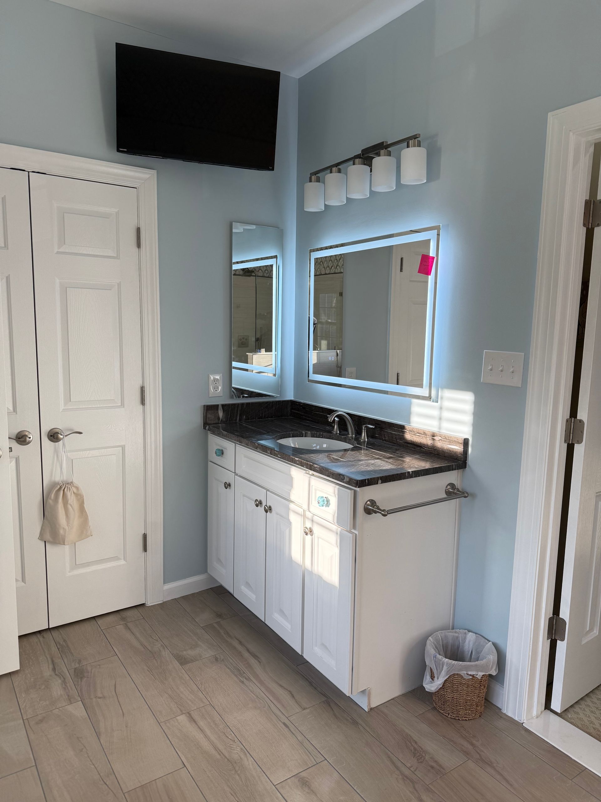 Bathroom vanity with mirror, black countertop, white cabinets, and wall-mounted TV in a light gray room