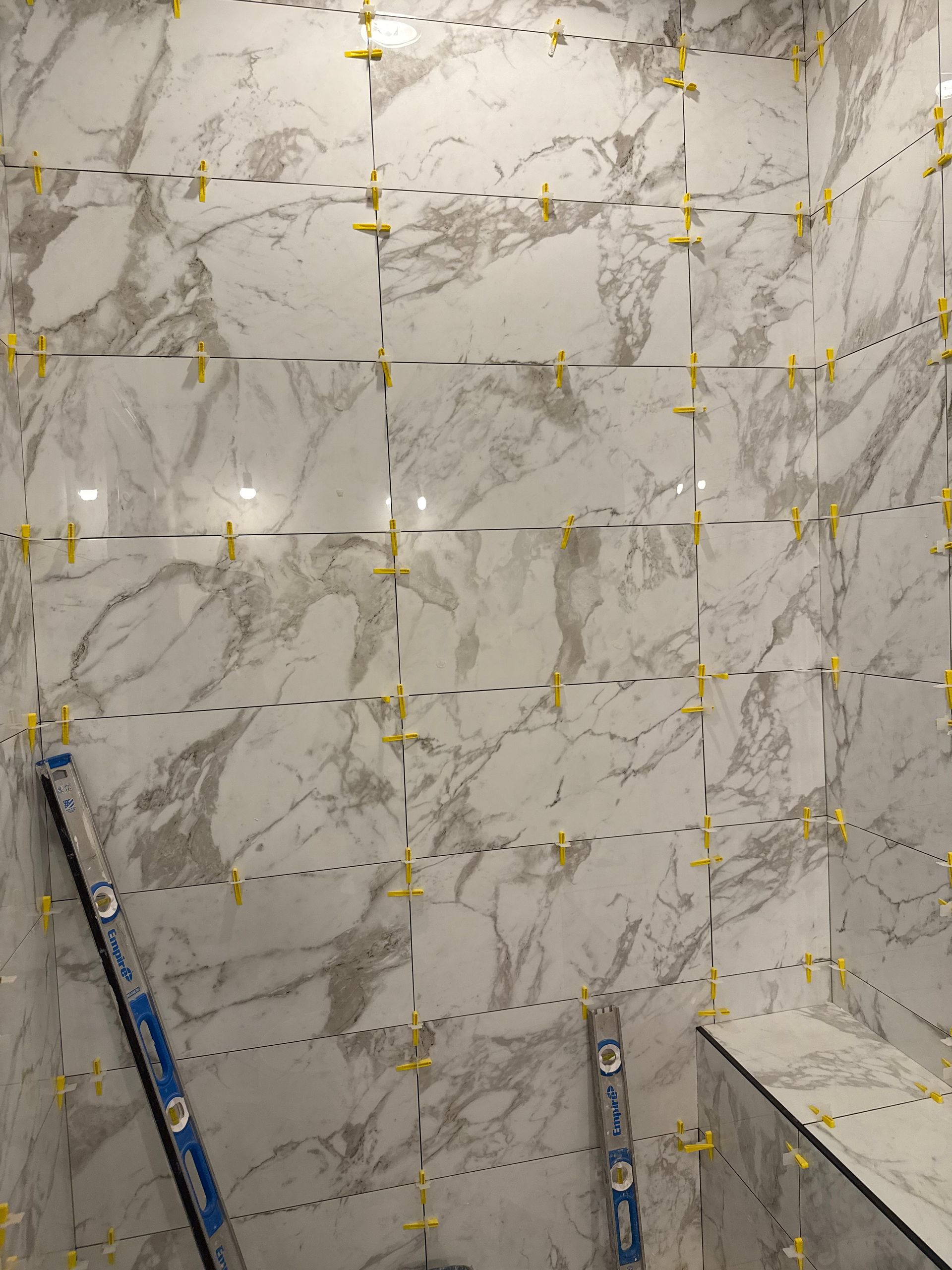 White marble-look tiled shower wall with gold tile spacers and blue tile tools leaning below