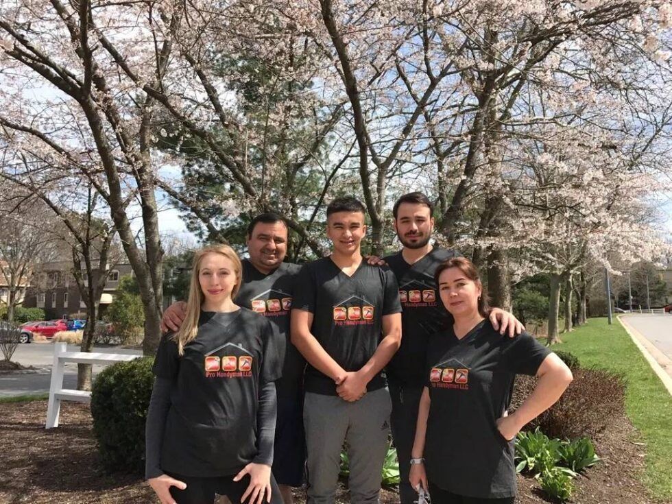 Four people in matching black shirts stand outdoors under blooming trees on a sunny day.
