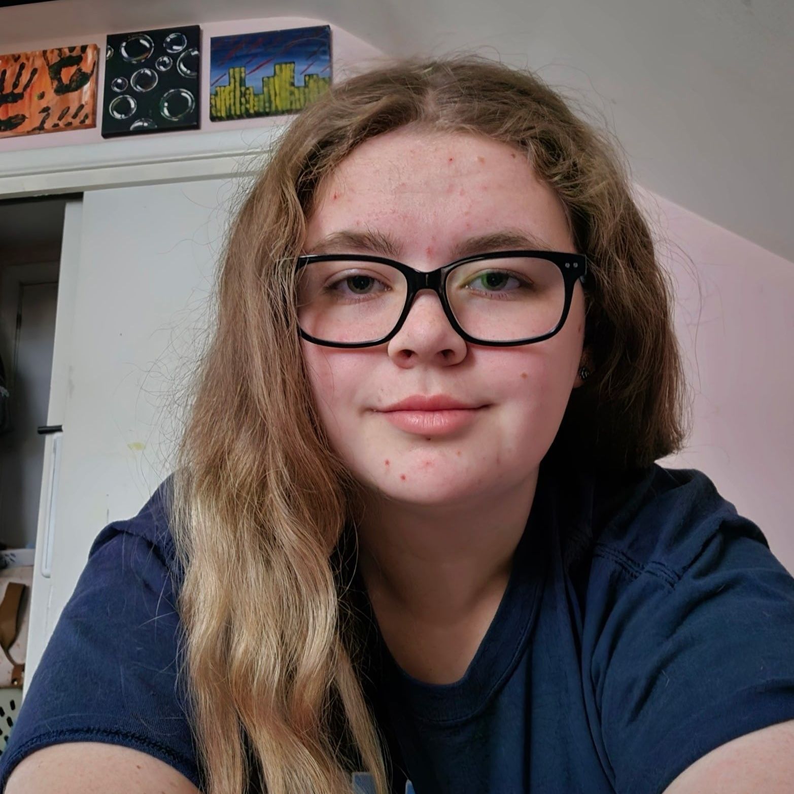 A young woman wearing glasses and a blue shirt is taking a selfie.