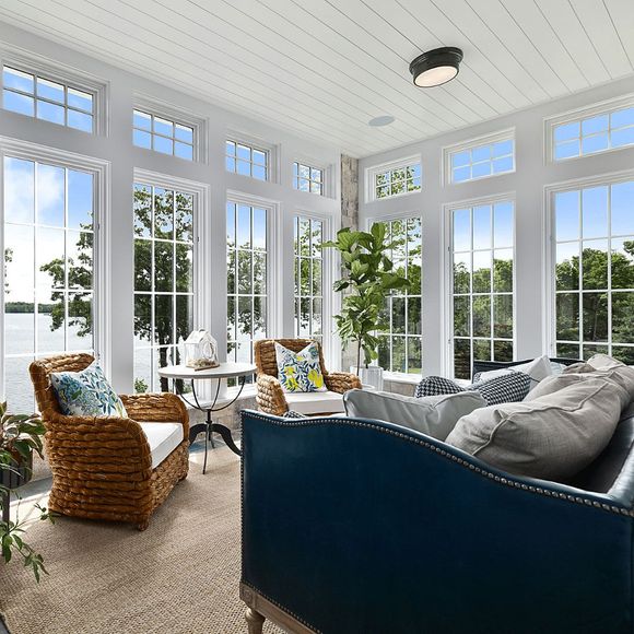 Sunroom with blue sofa and rattan chairs, overlooking a lake through large windows.