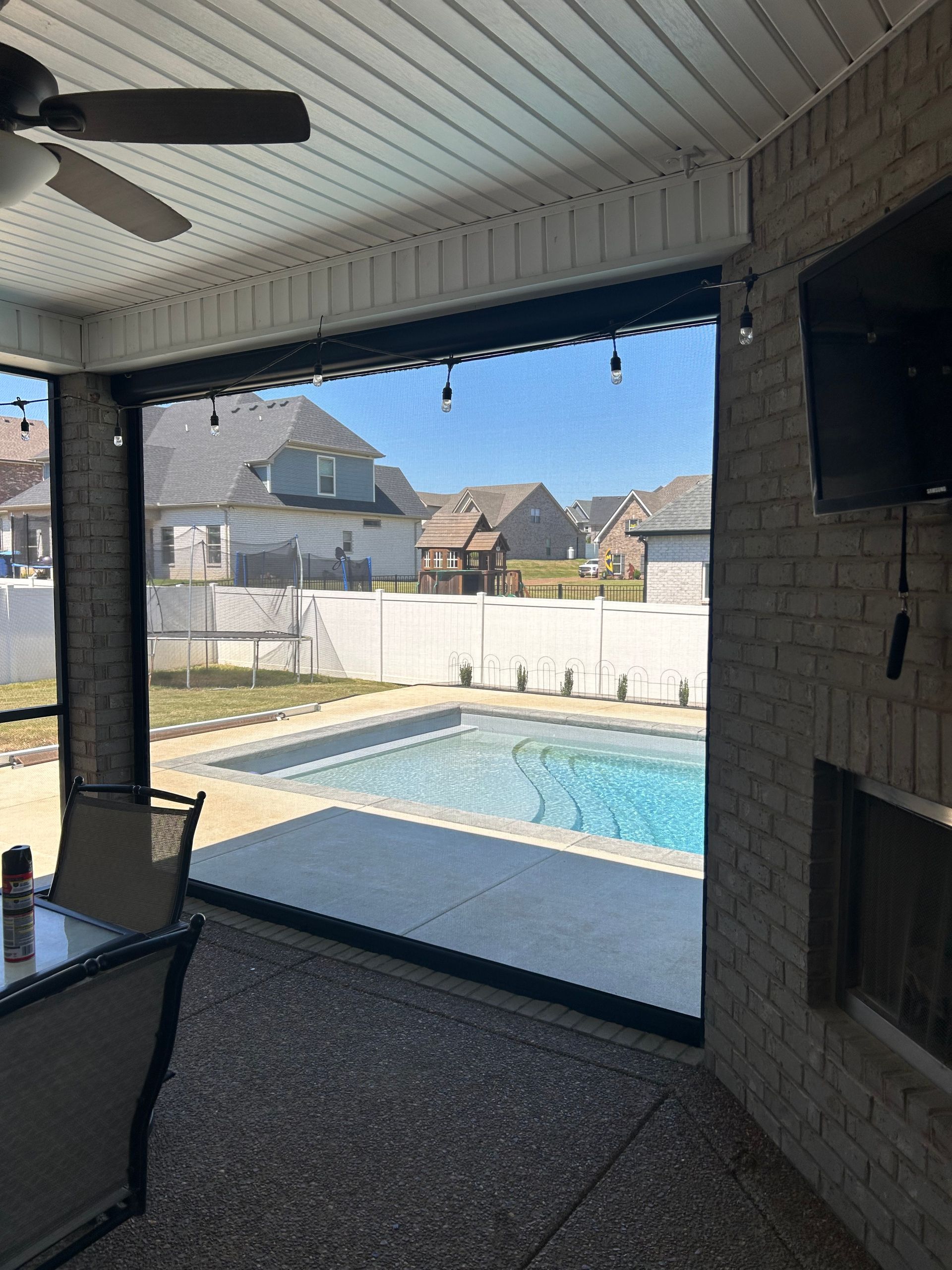 Covered patio with pool view; motorized screen shades, brick, ceiling fan, and outdoor TV.