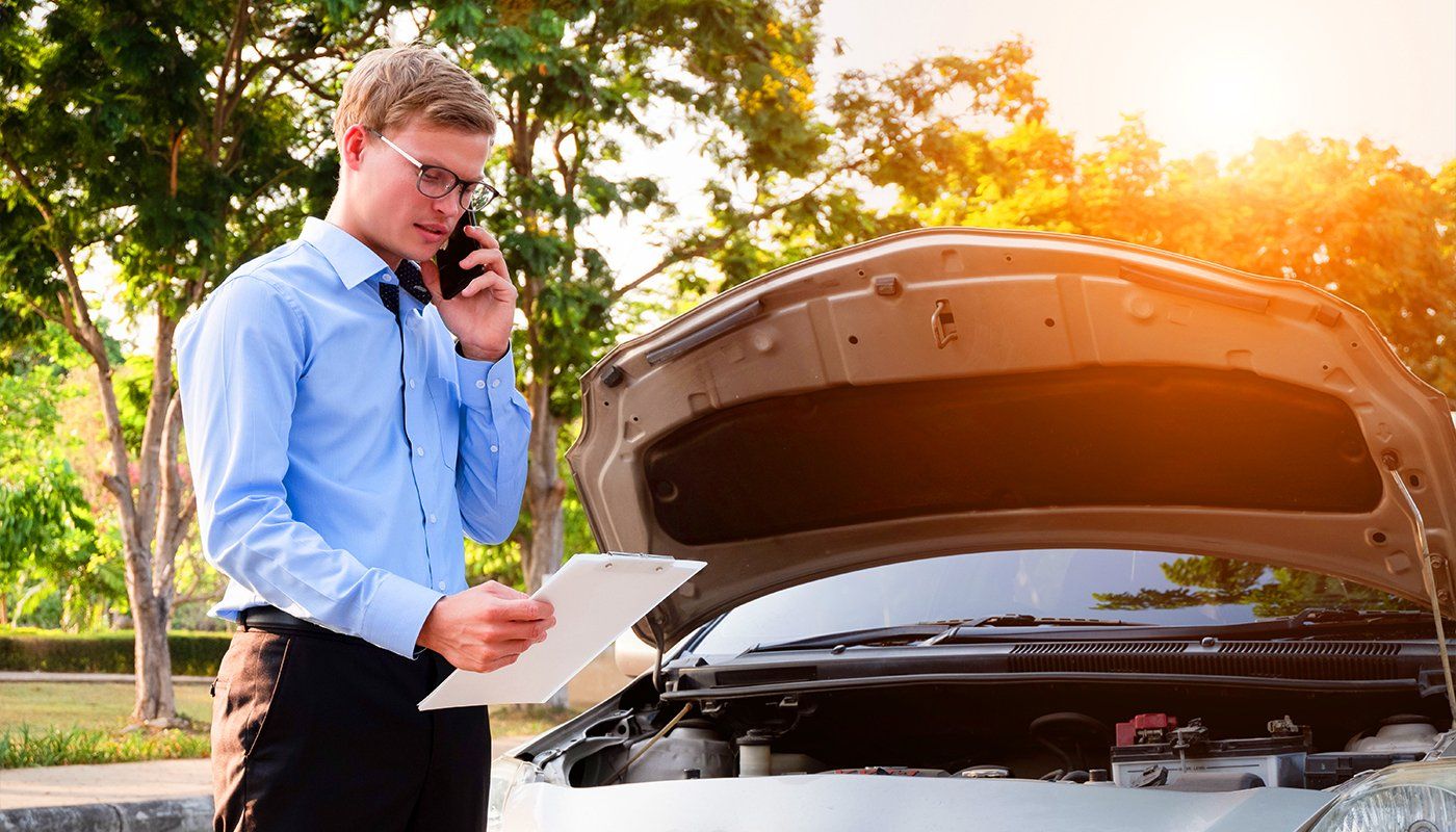 Learn More About Commercial Insurance A man is standing next to a broken down car talking on a cell phone