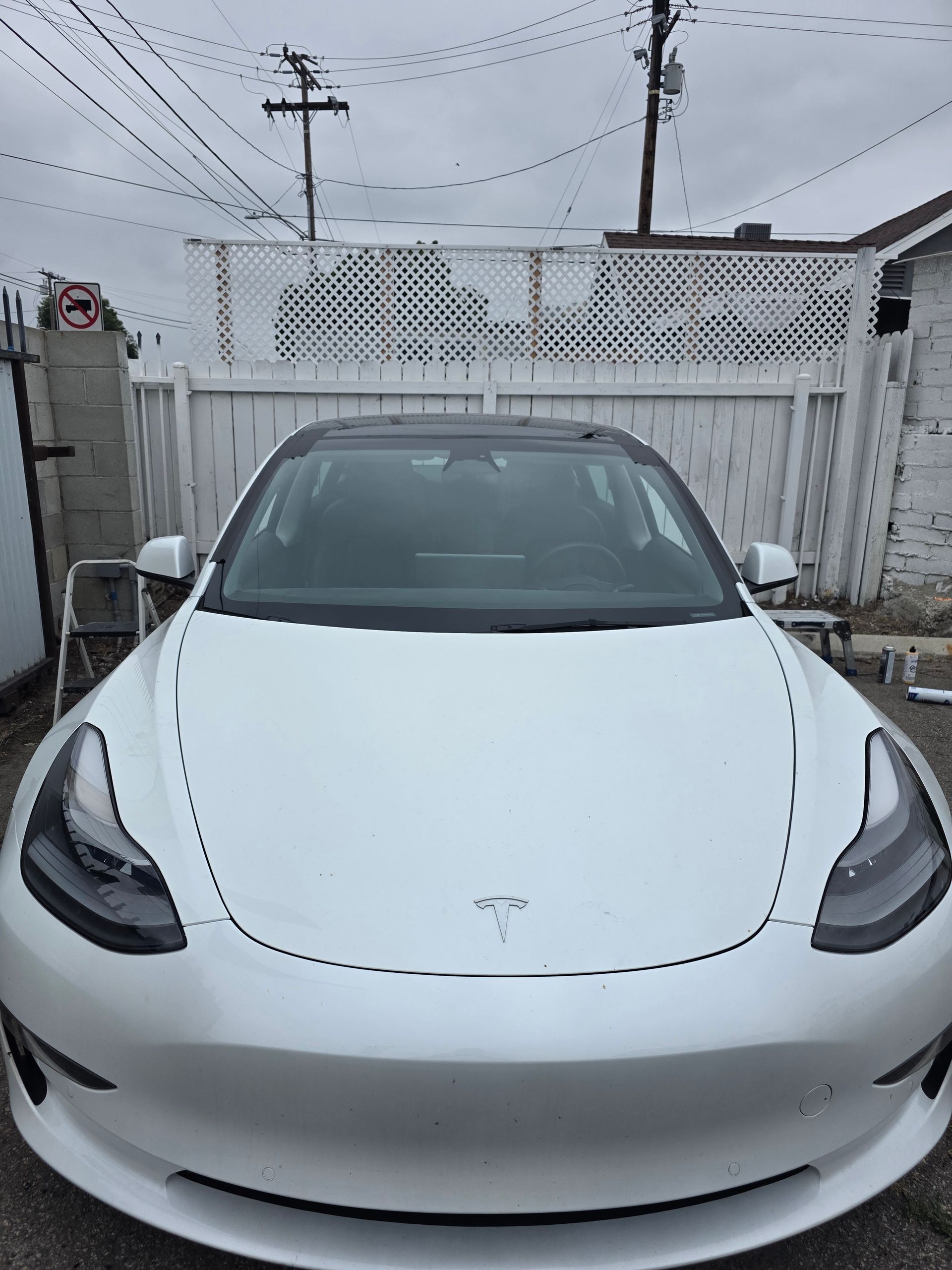 White Tesla parked in front of a white picket fence, with power lines overhead, under a cloudy sky.