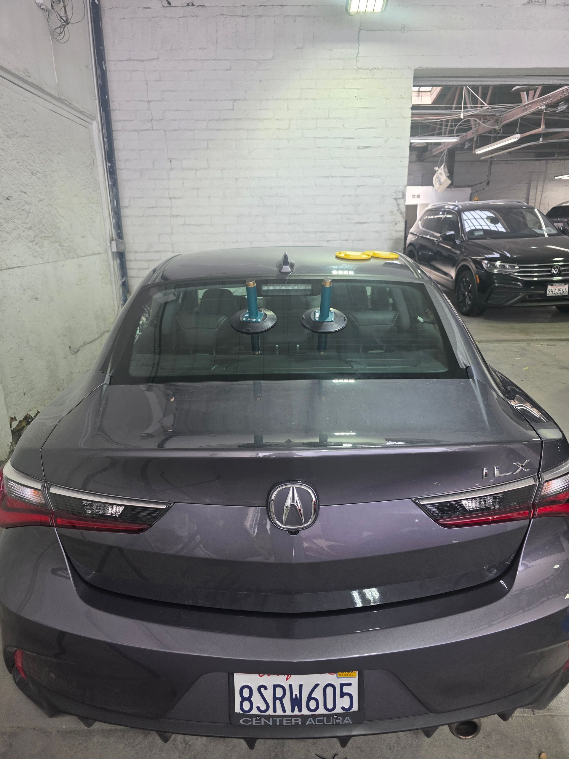 Rear view of a gray Acura sedan with suction cups attached to the rear window, likely for removal or replacement in a garage.