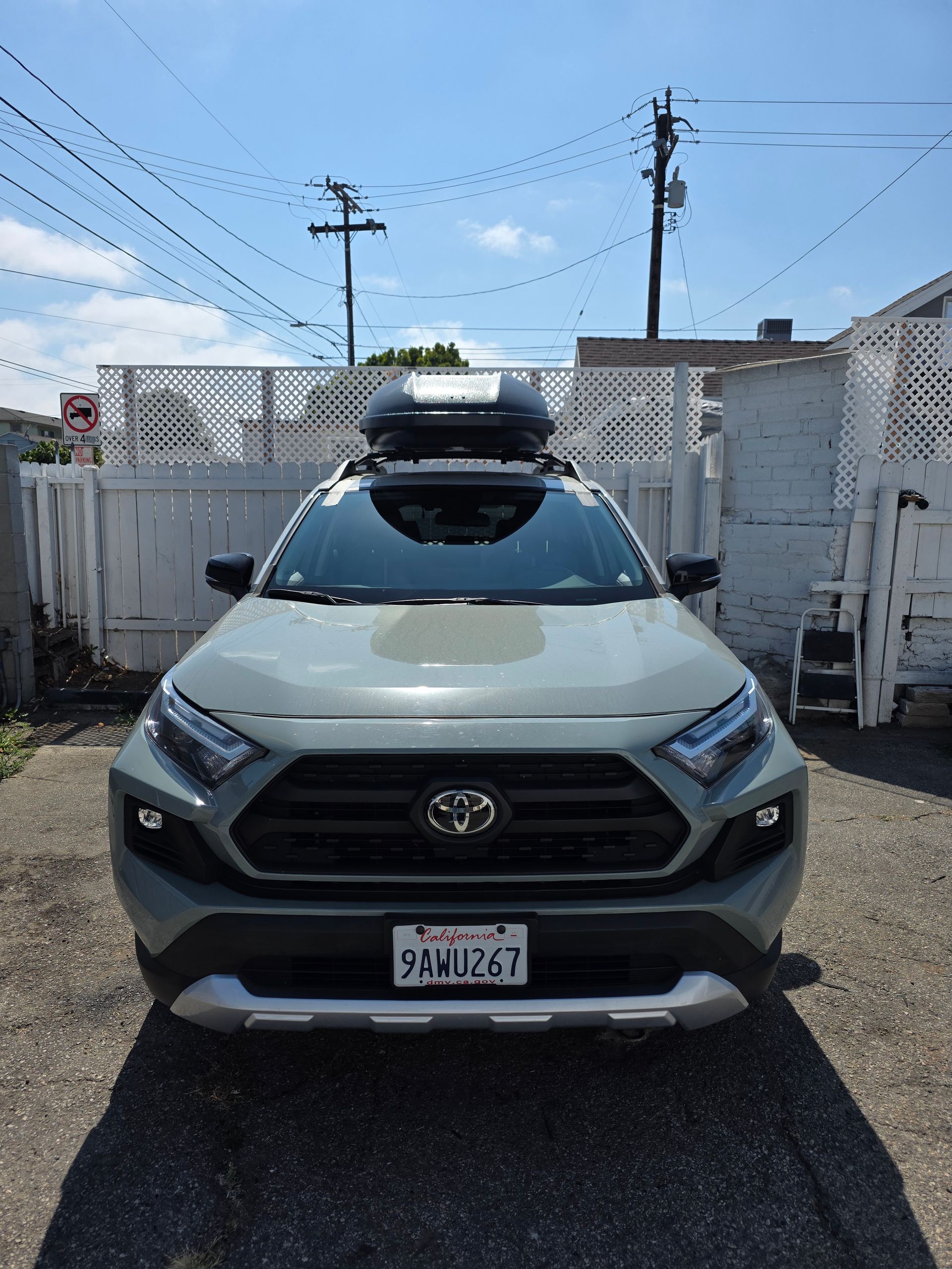 A gray Toyota RAV4 with a rooftop cargo carrier, parked in front of a white fence on a sunny day.