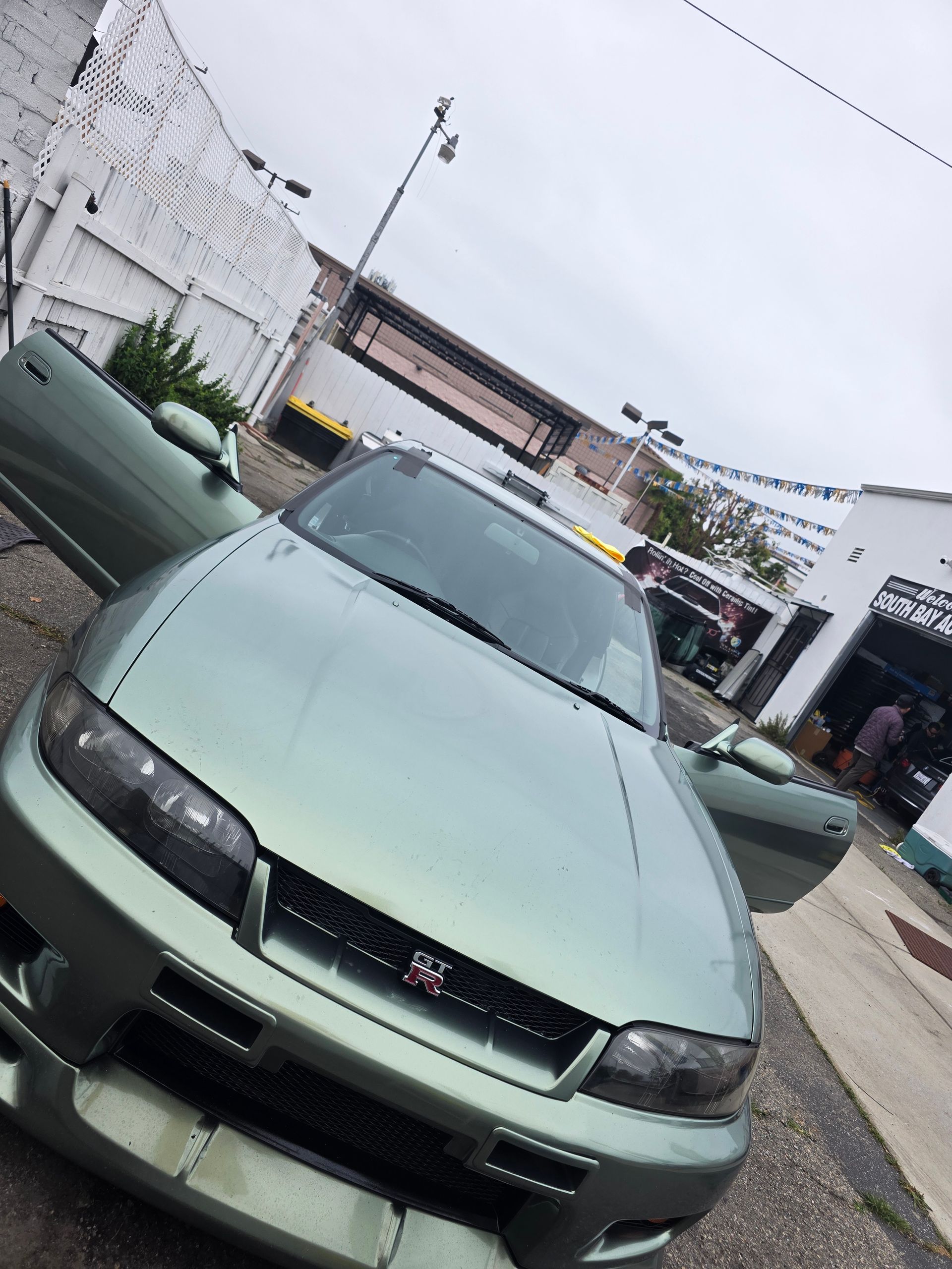 A green Nissan Skyline R33 with open doors parked on a street. The sky is overcast.