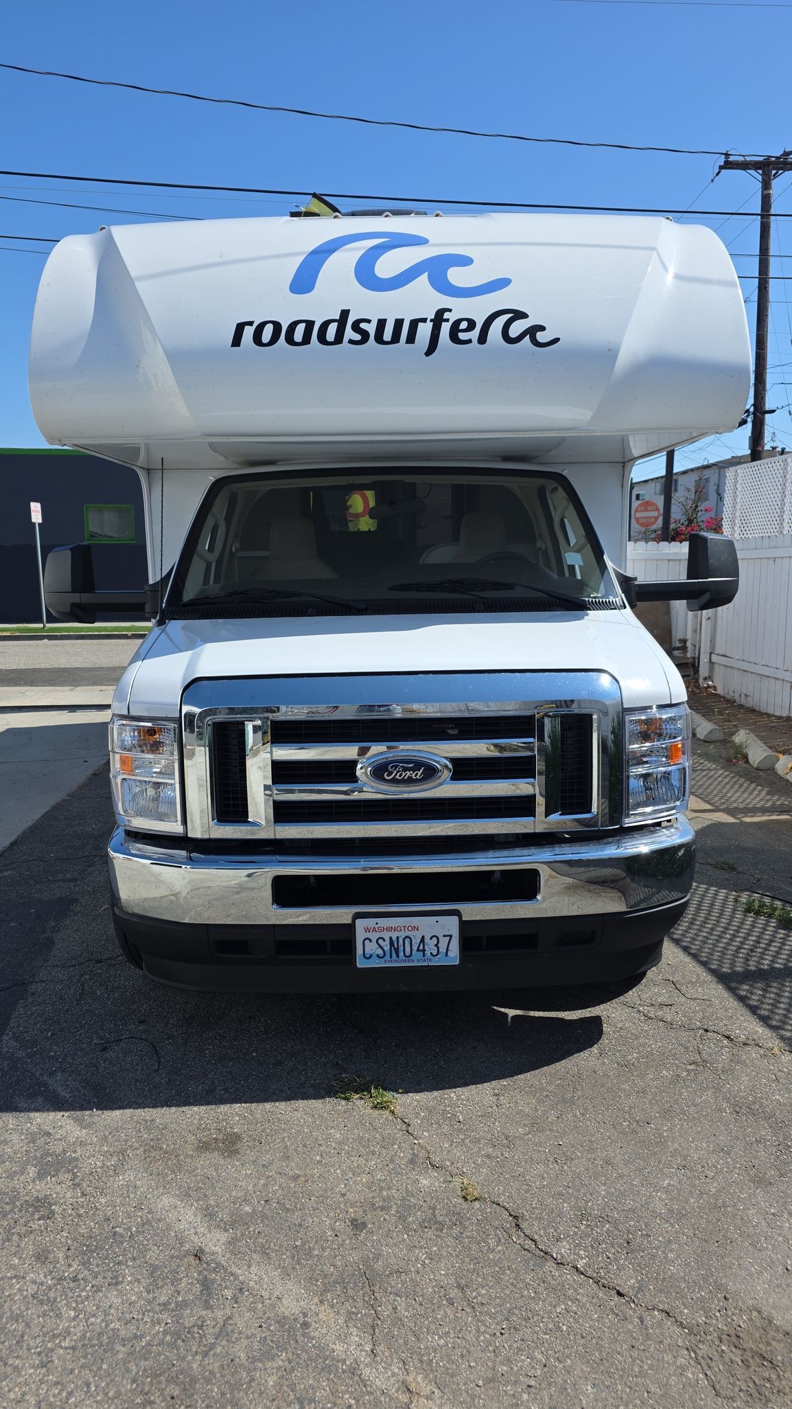 White Ford Roadsurfer RV parked on pavement, under a blue sky. The vehicle's front is facing the viewer.