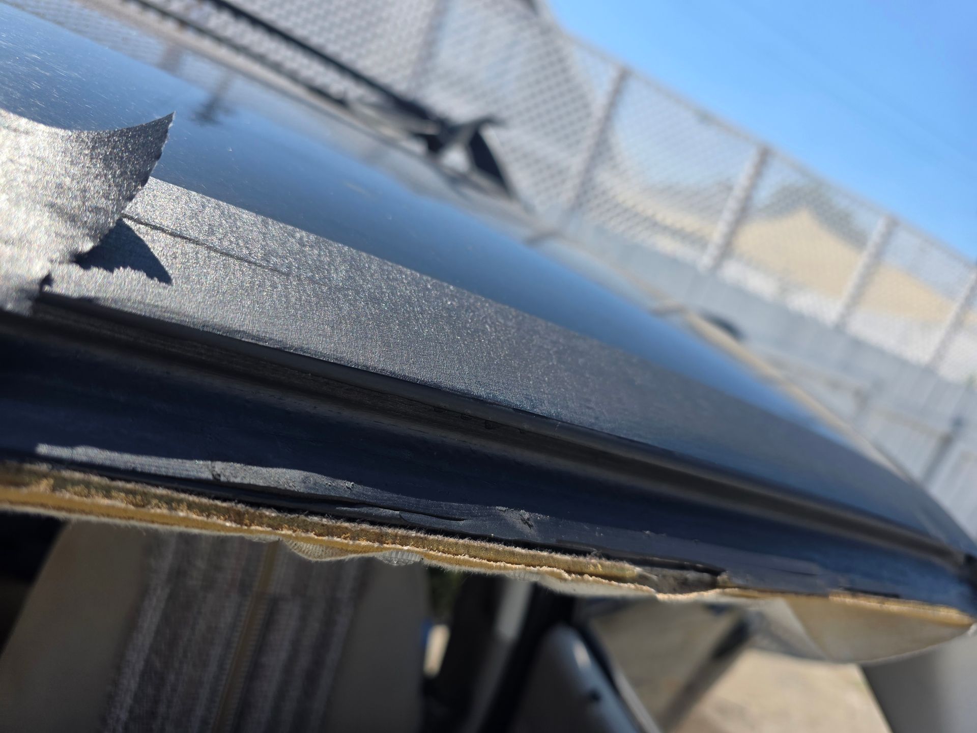 Close-up of a car roof, showing peeling black weather stripping. The setting appears to be outdoors, with a fence in the background.
