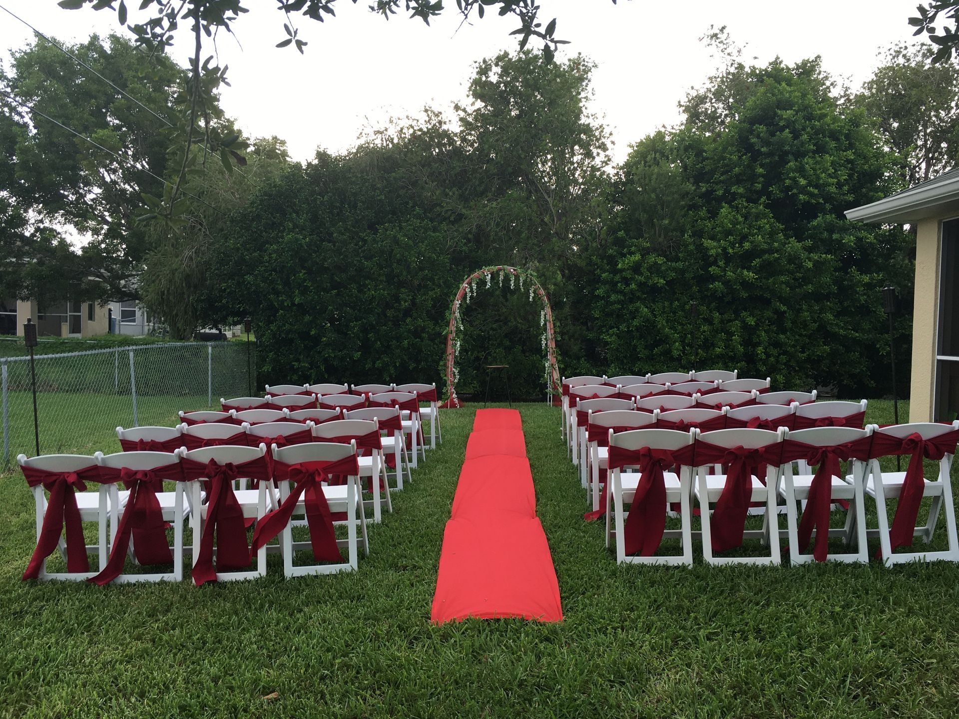 Outdoor wedding setup with white chairs and red sashes lining a red carpet leading to a floral arch