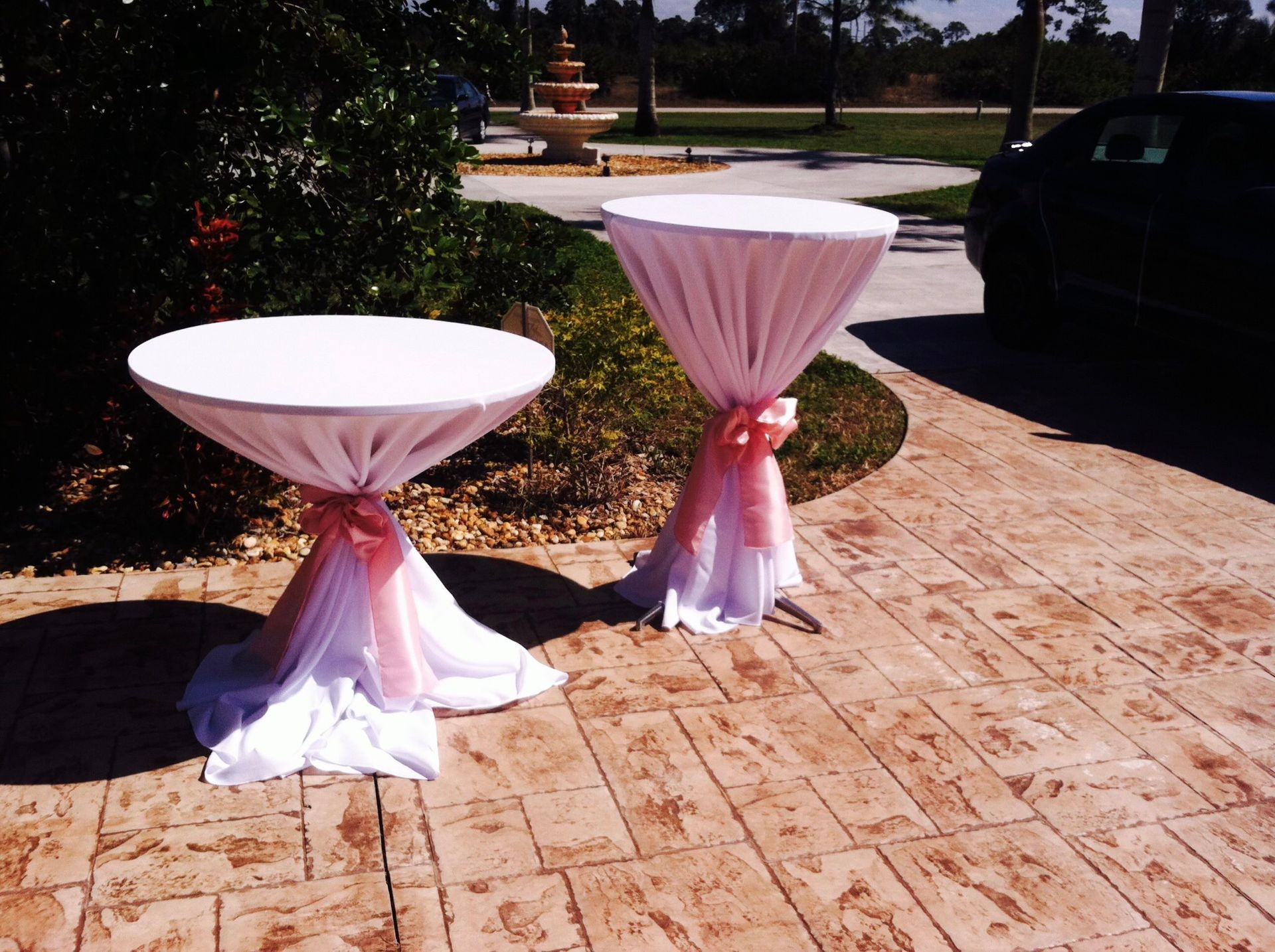 Two white cocktail tables, draped with white fabric and pink accents, sit outdoors on a brick patio