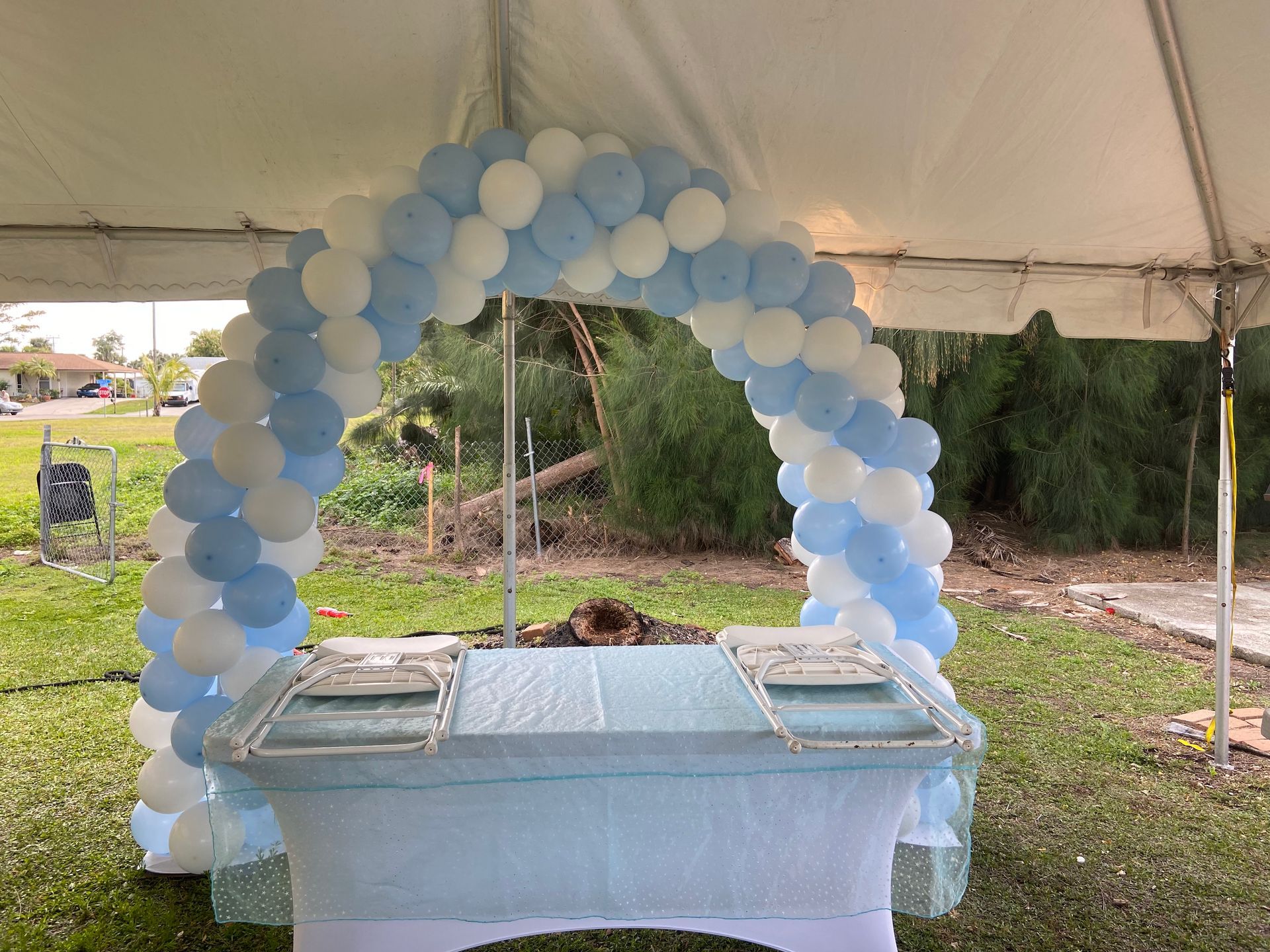 A light blue and white balloon arch frames a food serving table under a tent outdoors on a grassy lawn
