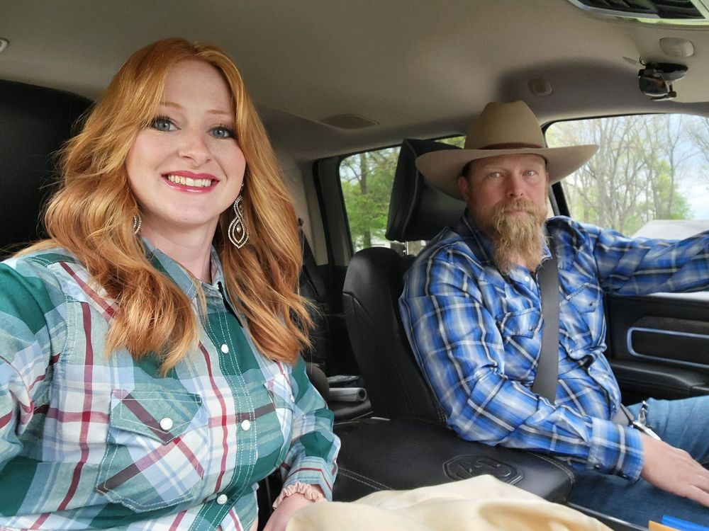 Woman with red hair and man with beard in truck, both smiling. Woman in green plaid shirt. Man wears cowboy hat and blue plaid.