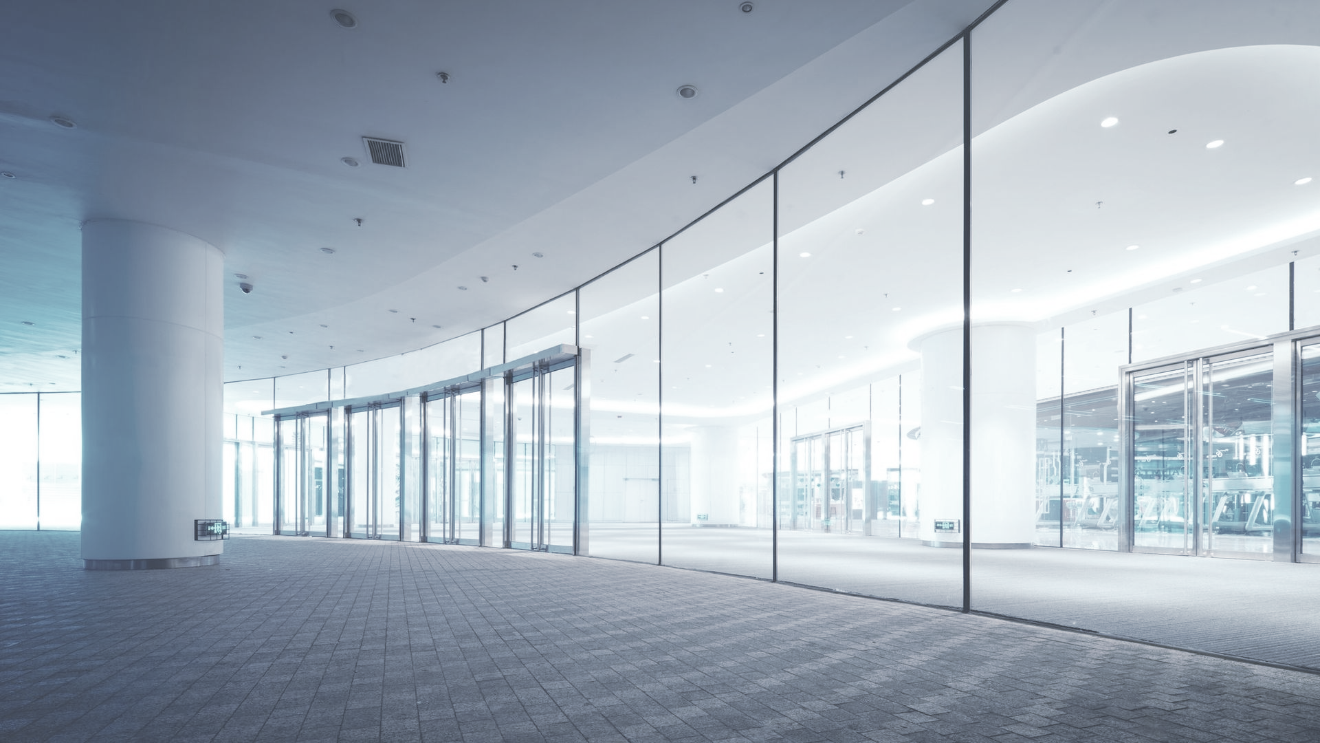 Large, empty lobby with curved glass wall, automatic doors, and white columns; carpeted floor.