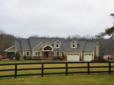 Green house with three garage doors, nestled in front of a treeline, behind a black wooden fence.