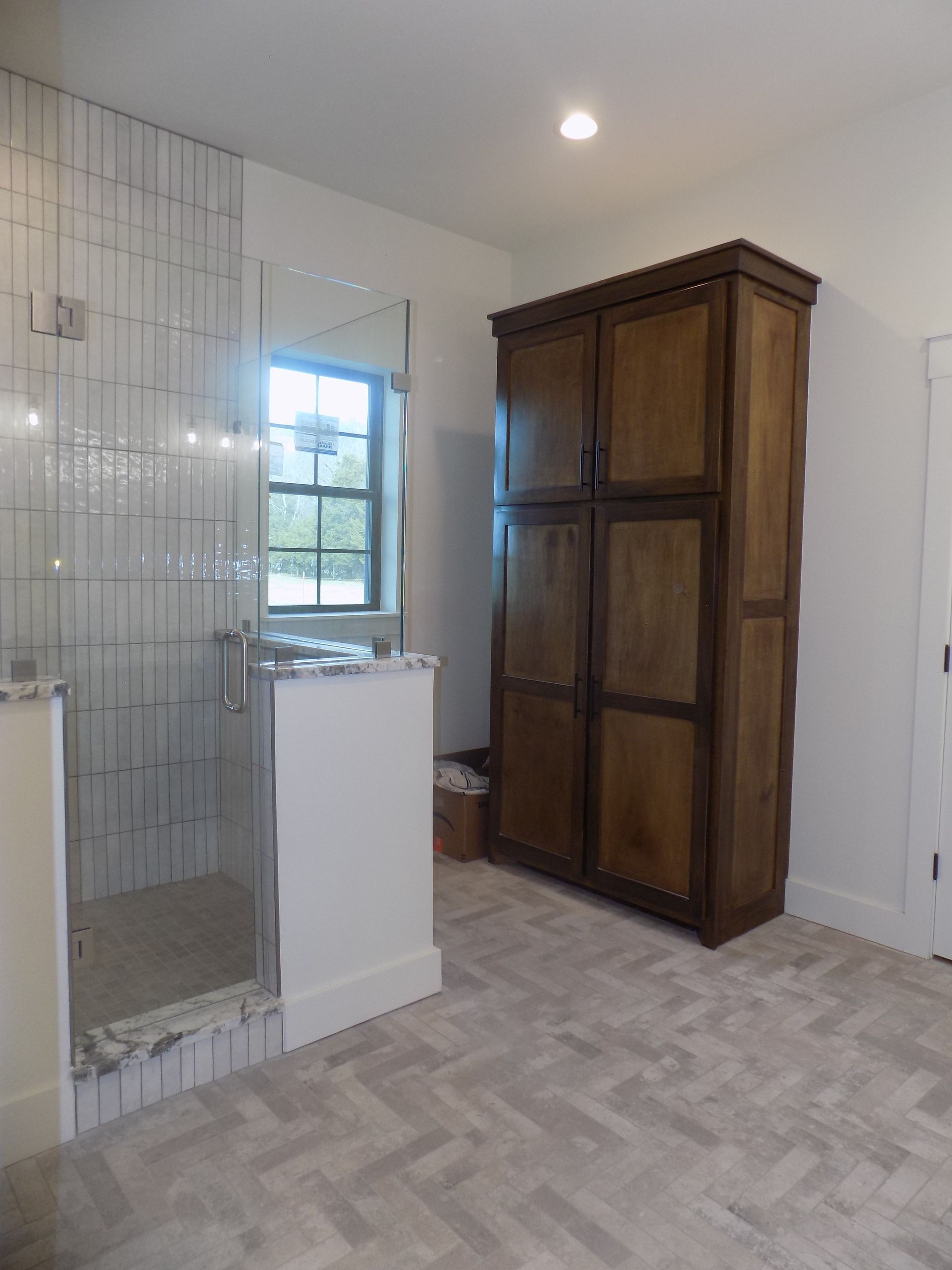 Bathroom with a tiled shower, wooden cabinet, and herringbone floor. A window is in the background.