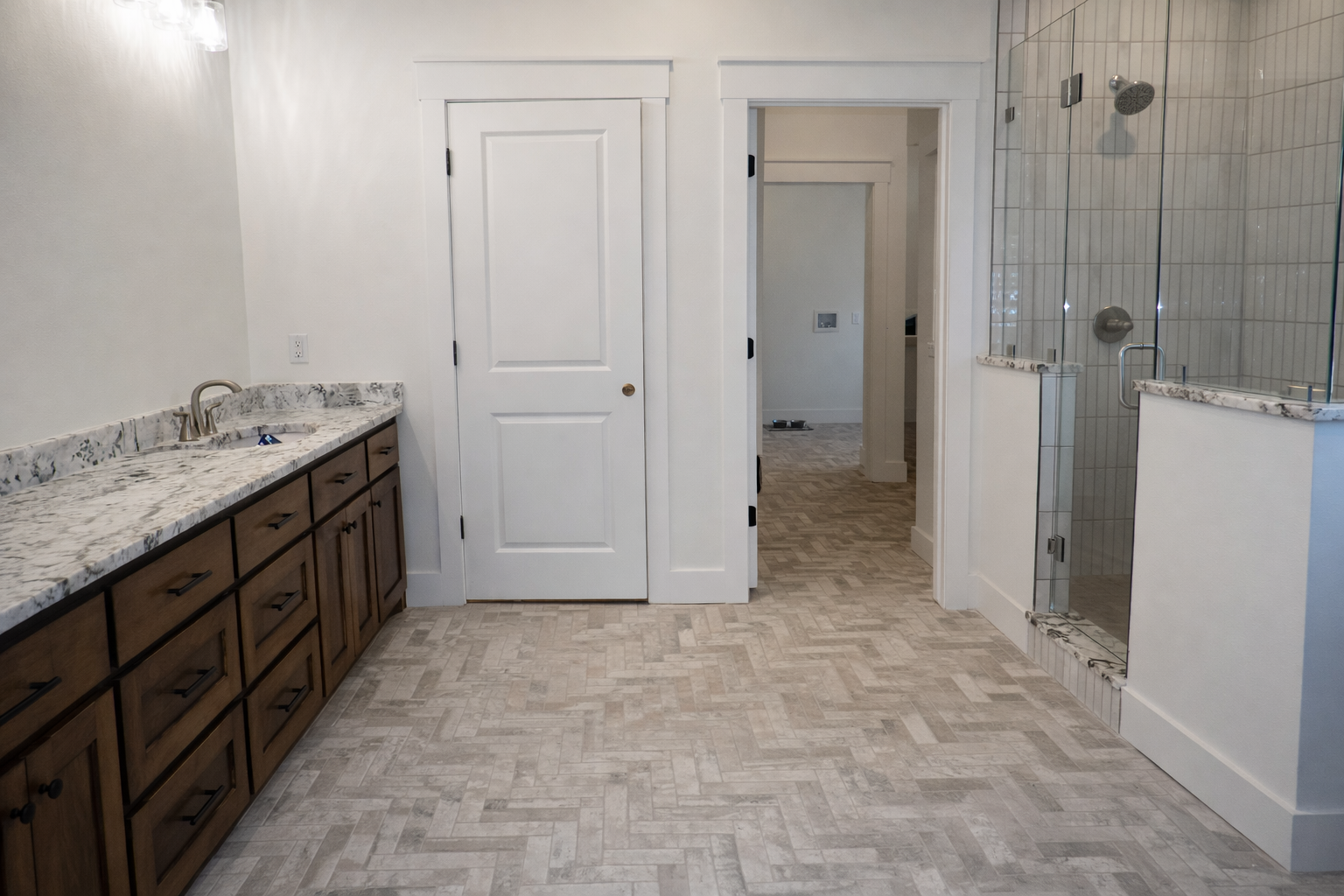 Bathroom with brown vanity, white door, and tiled floor, leading to shower and hallway.