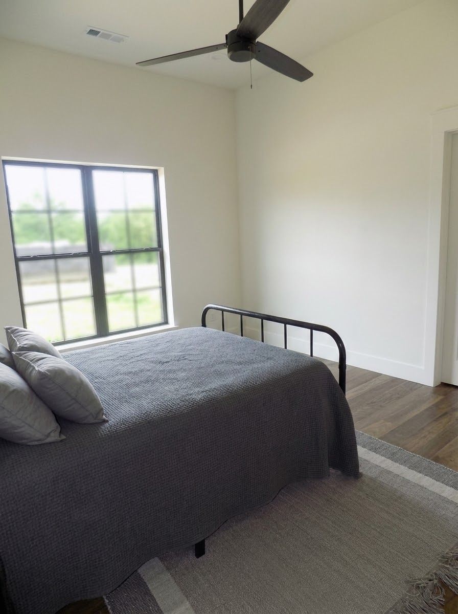 Bedroom with bed, window, and ceiling fan. Gray bedding, light walls, wooden floor, and rug.