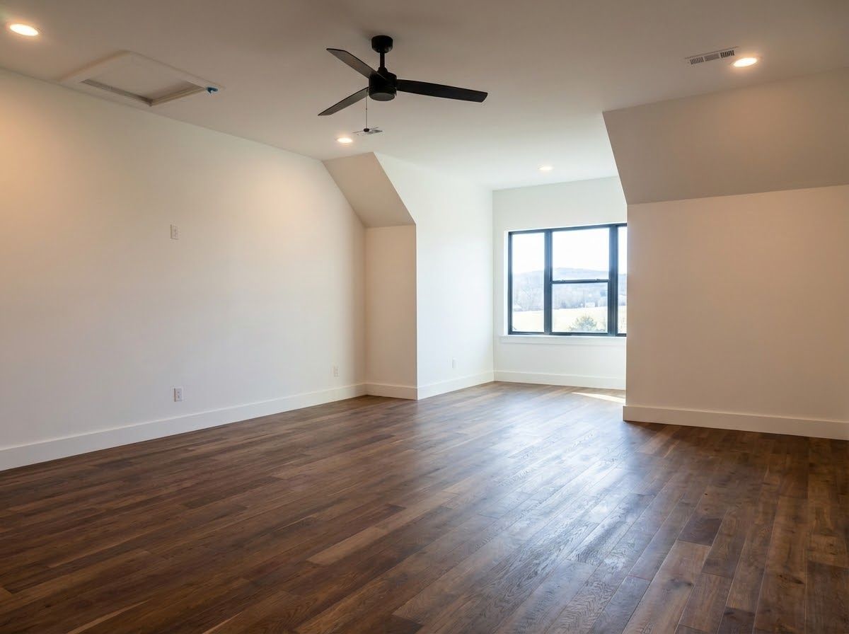 Empty room with dark wood floor, white walls, and a black ceiling fan. Window and attic access present.