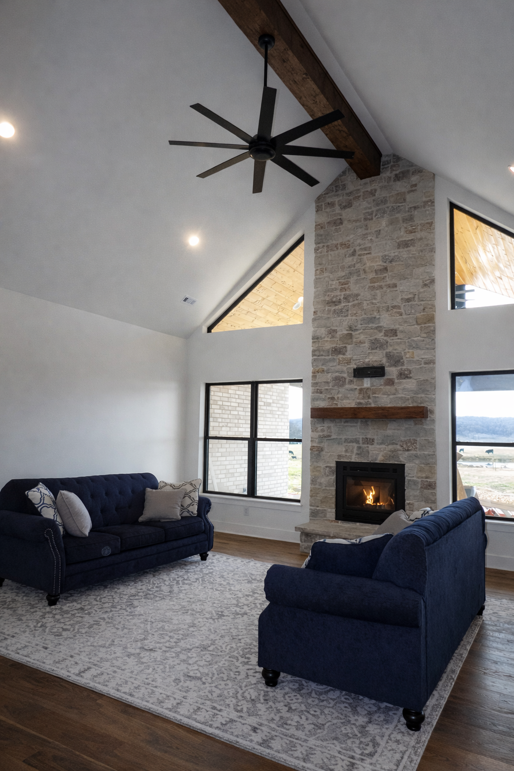 Living room with a stone fireplace, dark blue sofas, and a large black ceiling fan.