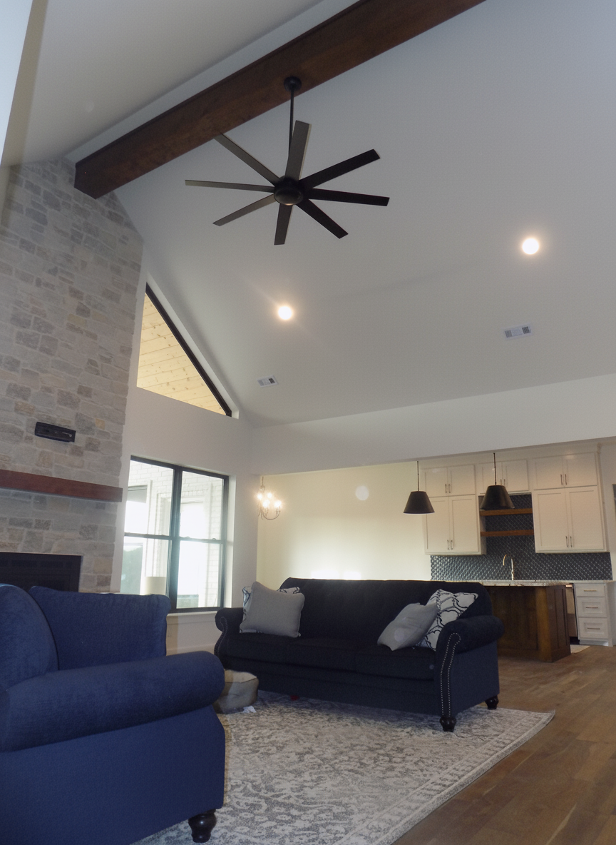 Living room with high ceiling, brick fireplace, dark sofa, and a ceiling fan.