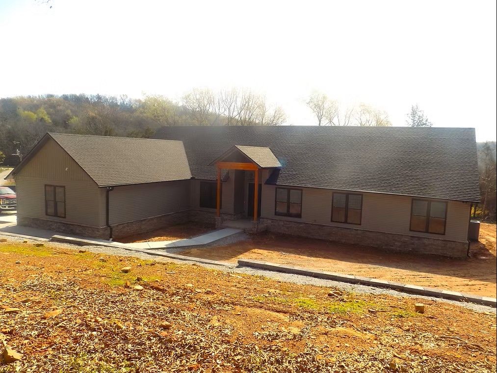 A single-story house with a gray exterior, brown roof, and stone accents. The house is on a slightly sloped lot.