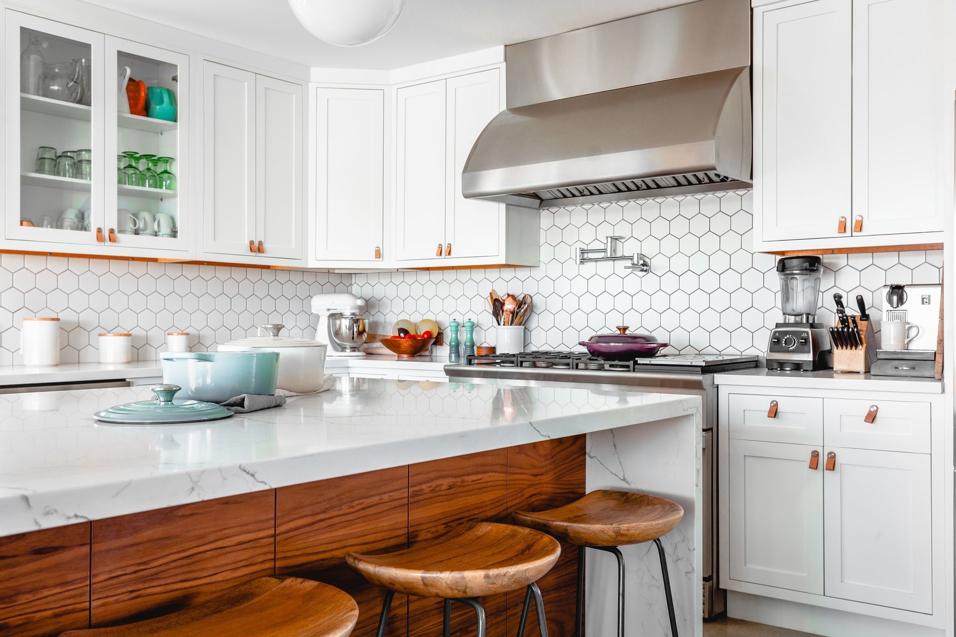 Modern white kitchen with island, white cabinets, honeycomb backsplash, stainless steel hood, and wood stools.